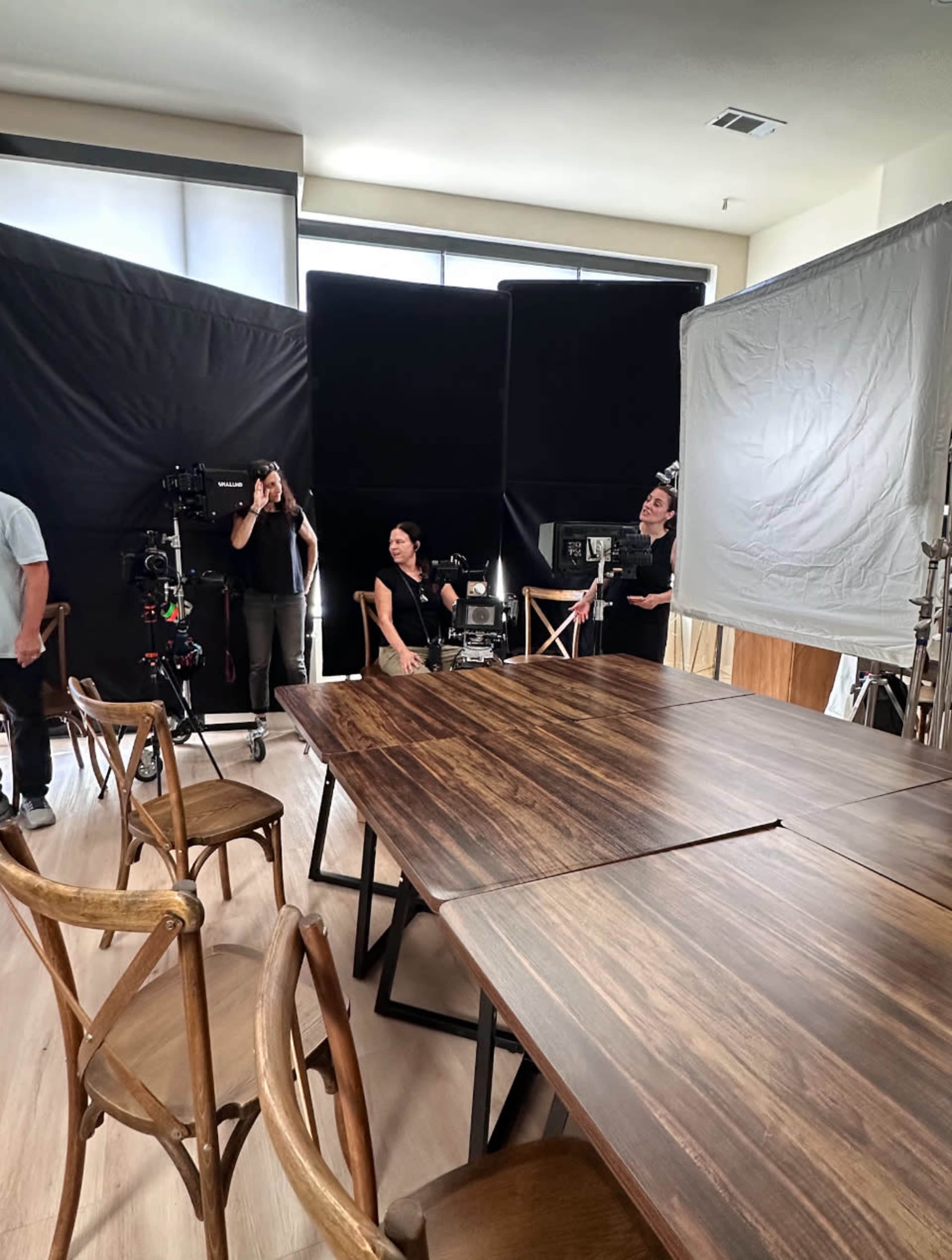 A film crew sets up equipment in a spacious room with a large wooden table and several chairs.