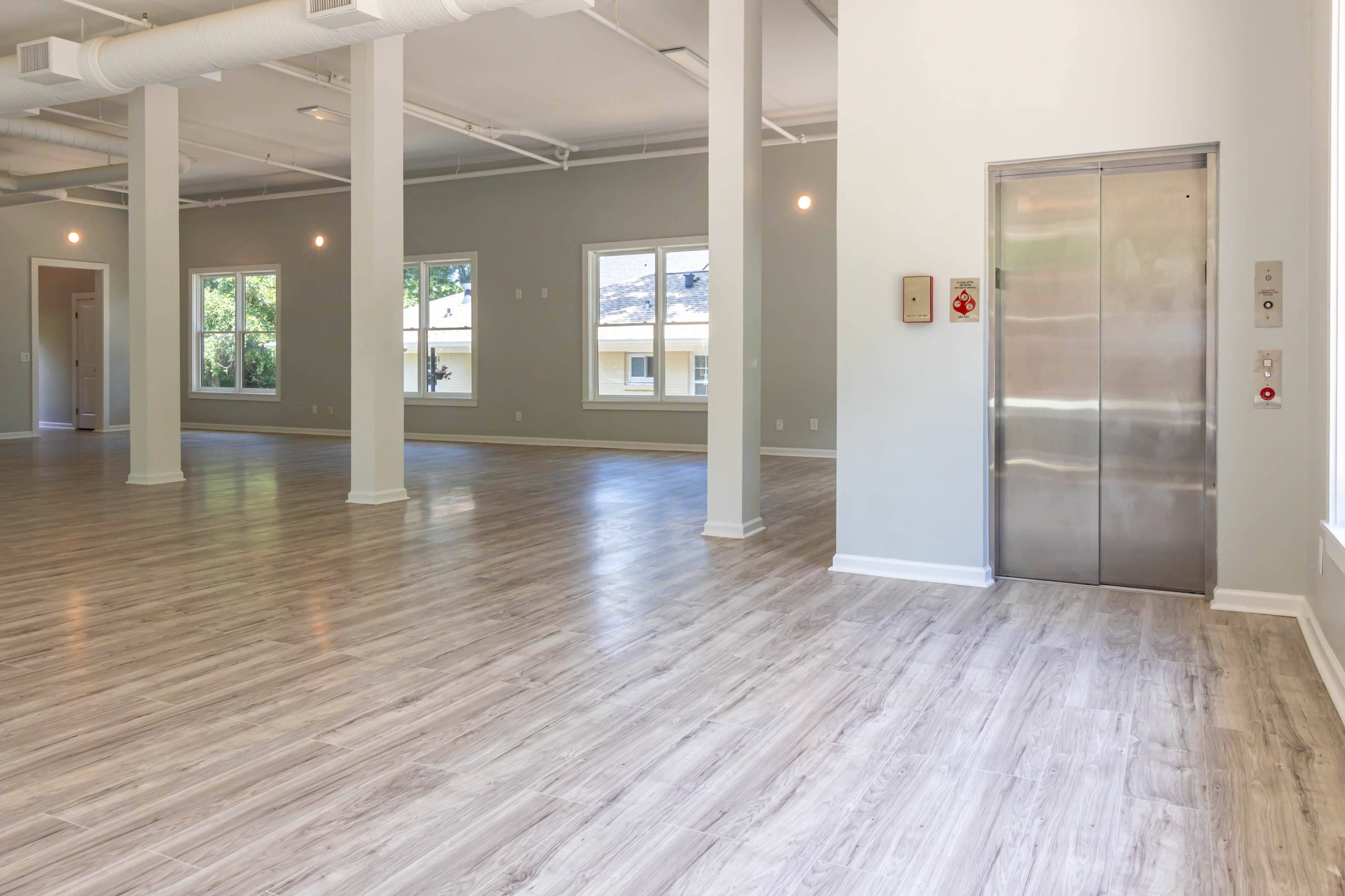 The image shows a spacious, well-lit interior with wooden floors and an elevator at one end, flanked by large windows.