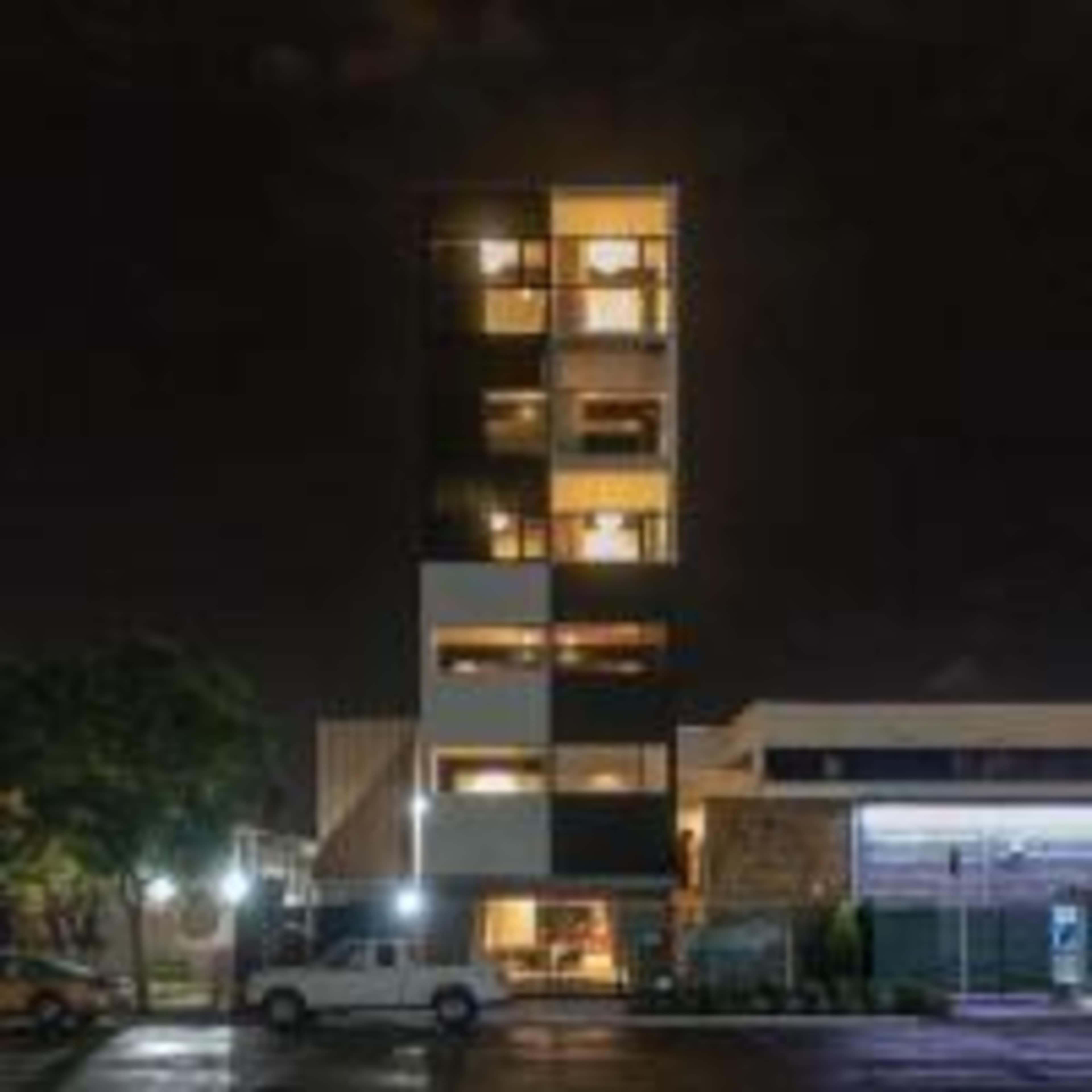 A tall, modern building with multiple illuminated balconies stands at night next to a parking area.