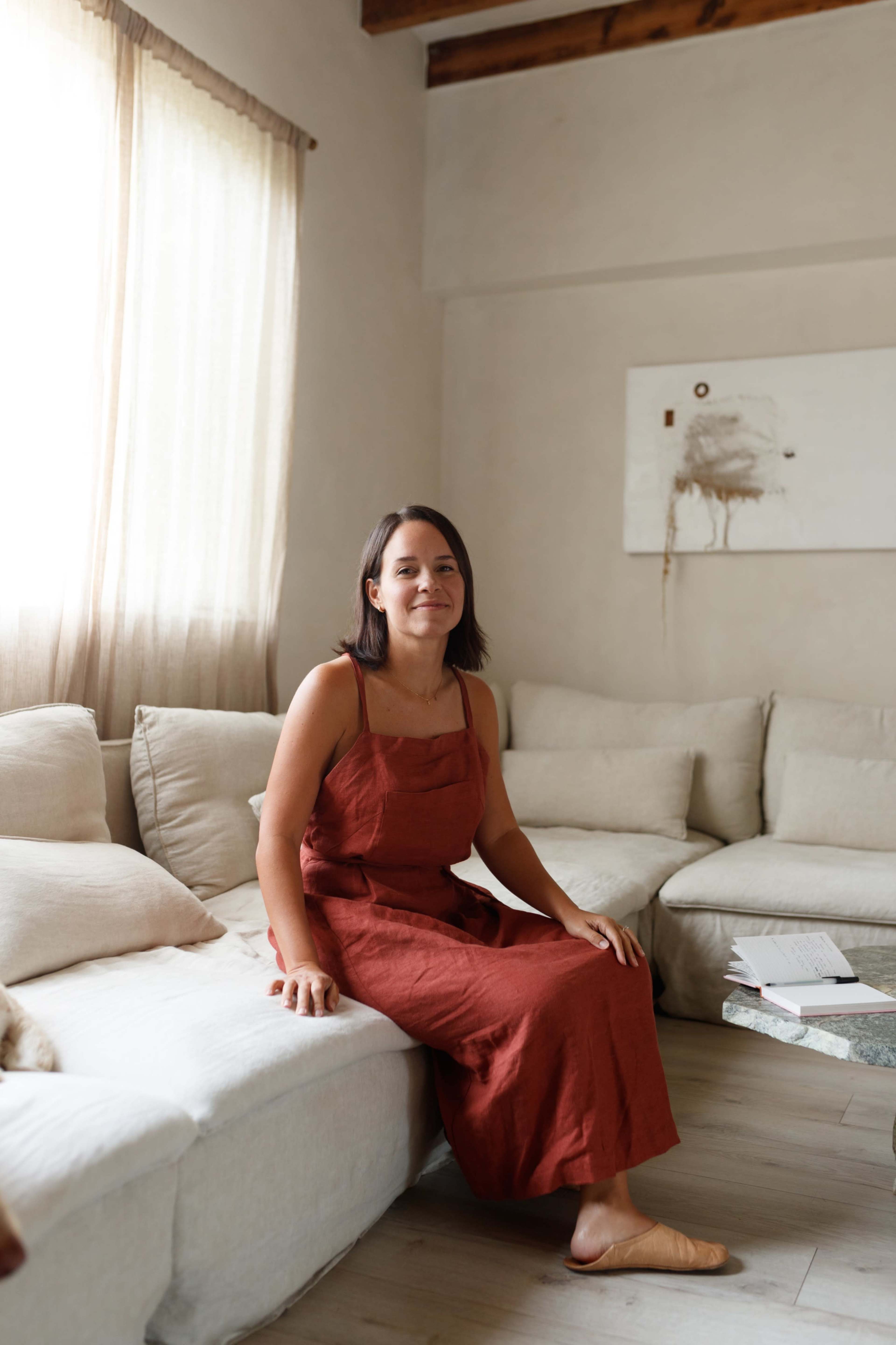 A woman sits on a light-colored sofa in a well-lit living room adorned with soft textiles and minimal artwork.