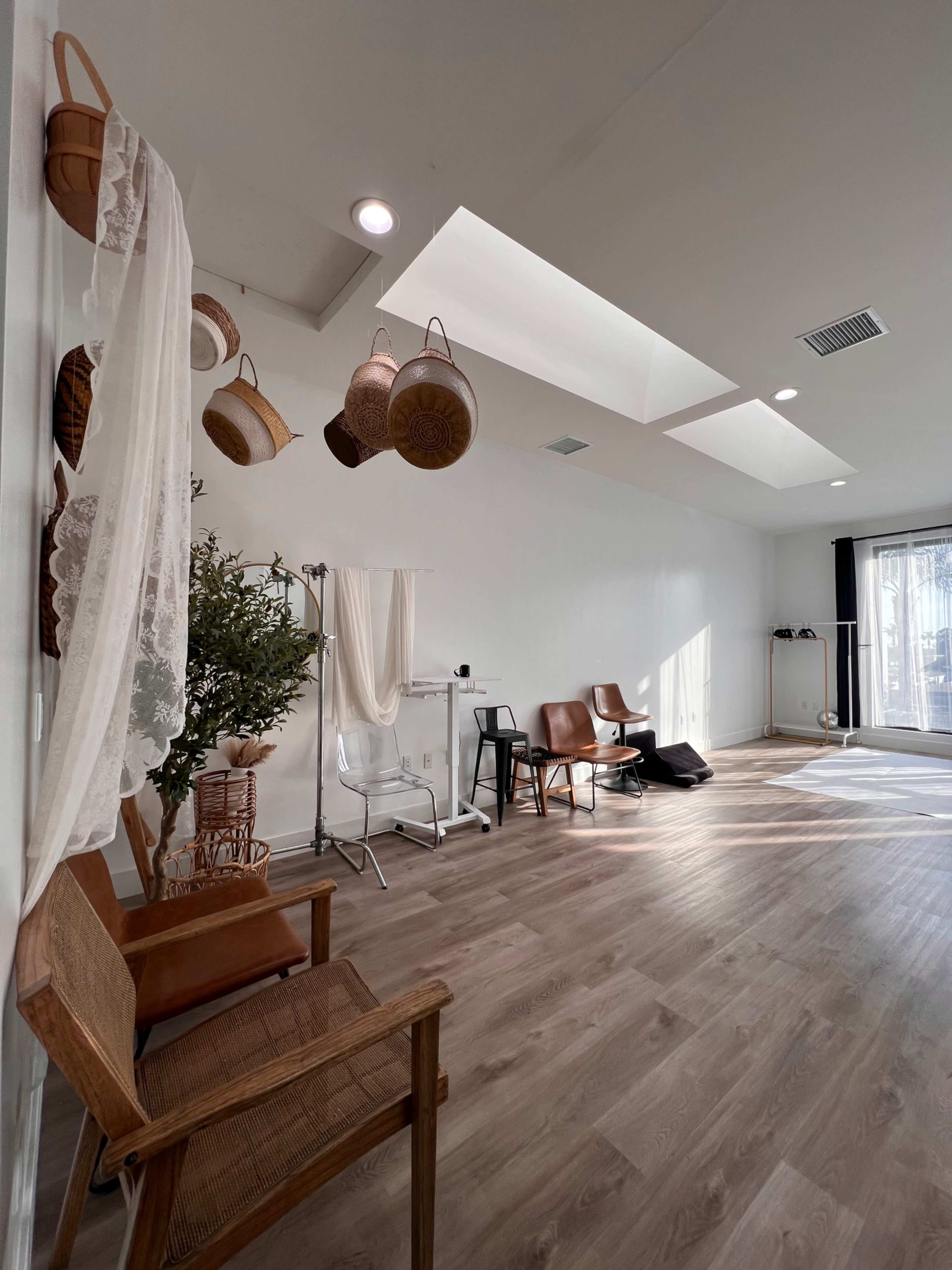 The image shows a spacious, well-lit room with wooden flooring, featuring chairs, a clothing rack, and baskets hanging on the wall.