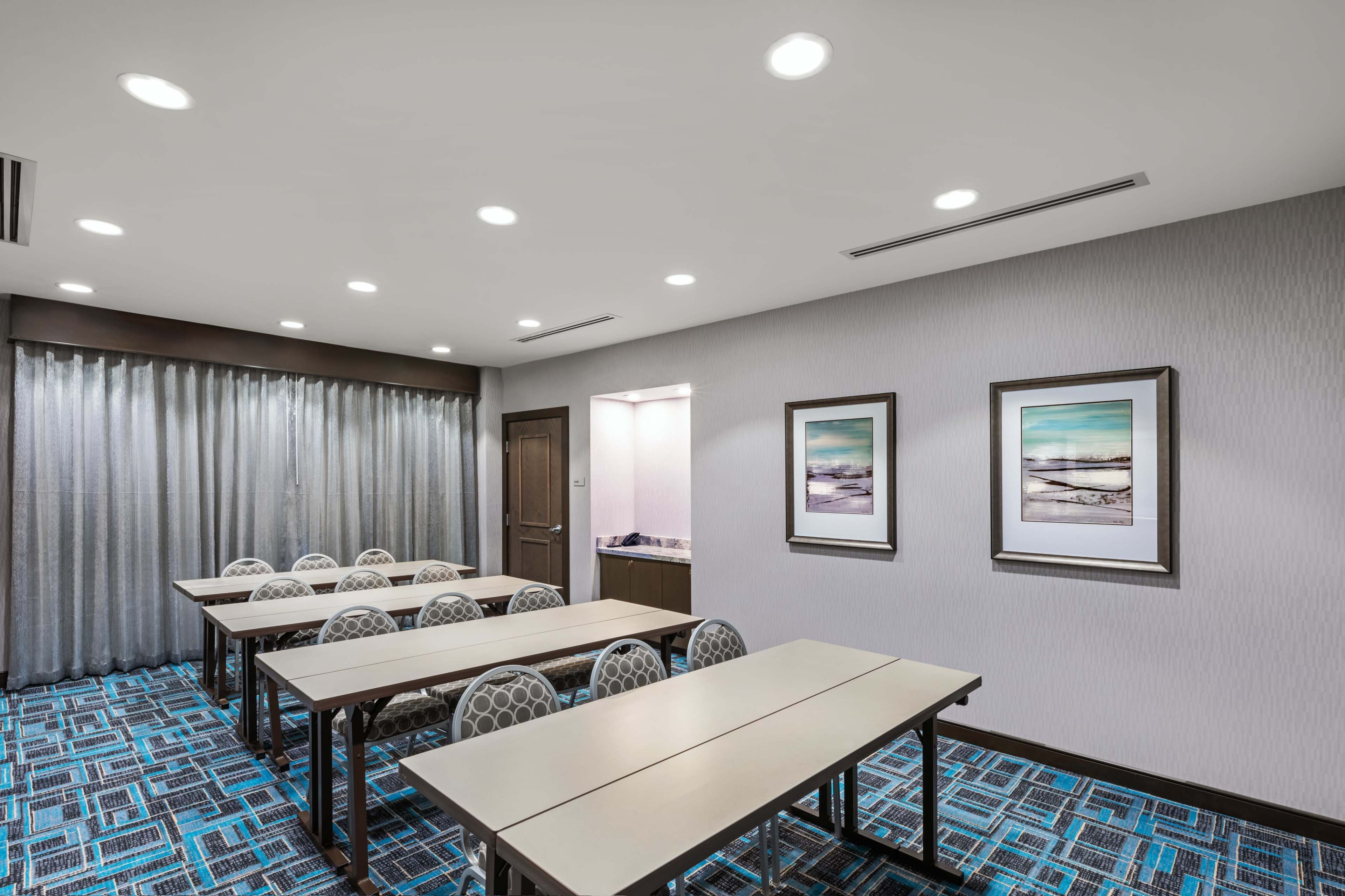 A meeting room features several arranged tables with chairs around them, alongside large windows covered with curtains, and two framed artworks on the walls.