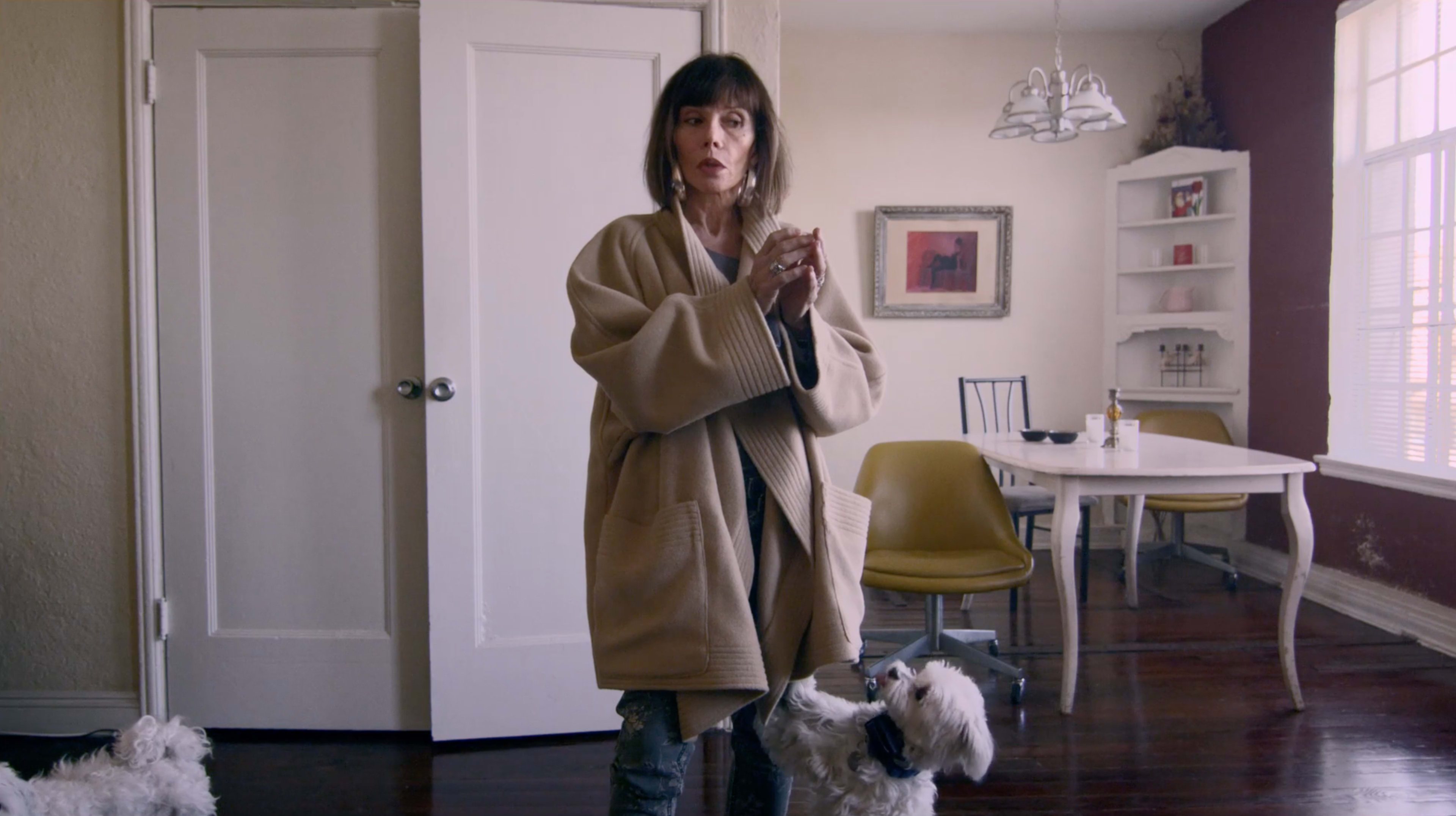 A woman in a beige robe stands in a kitchen while two small dogs are close by.