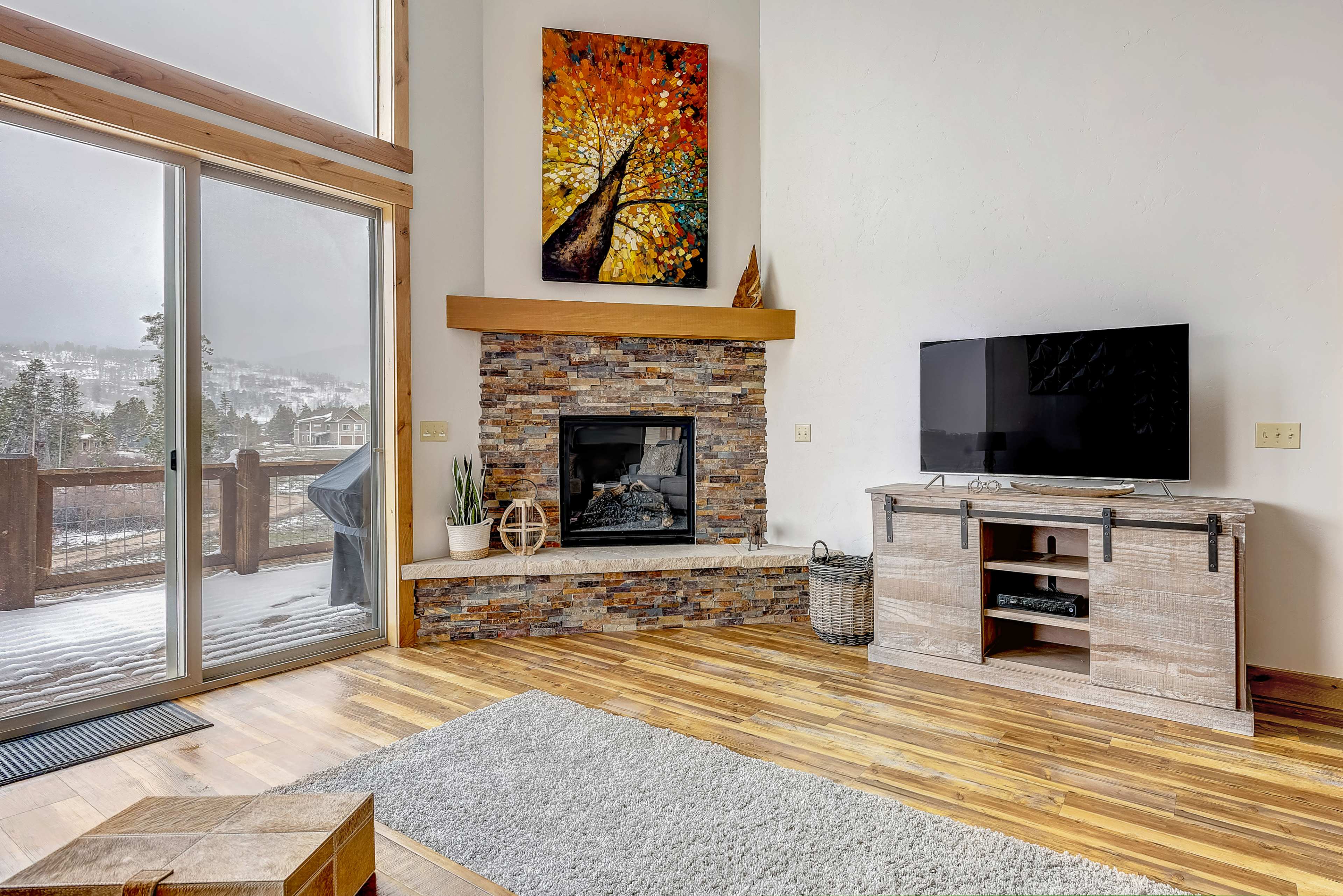 A spacious living room features a stone fireplace, a large television on a wooden cabinet, and a large window overlooking a snowy landscape.