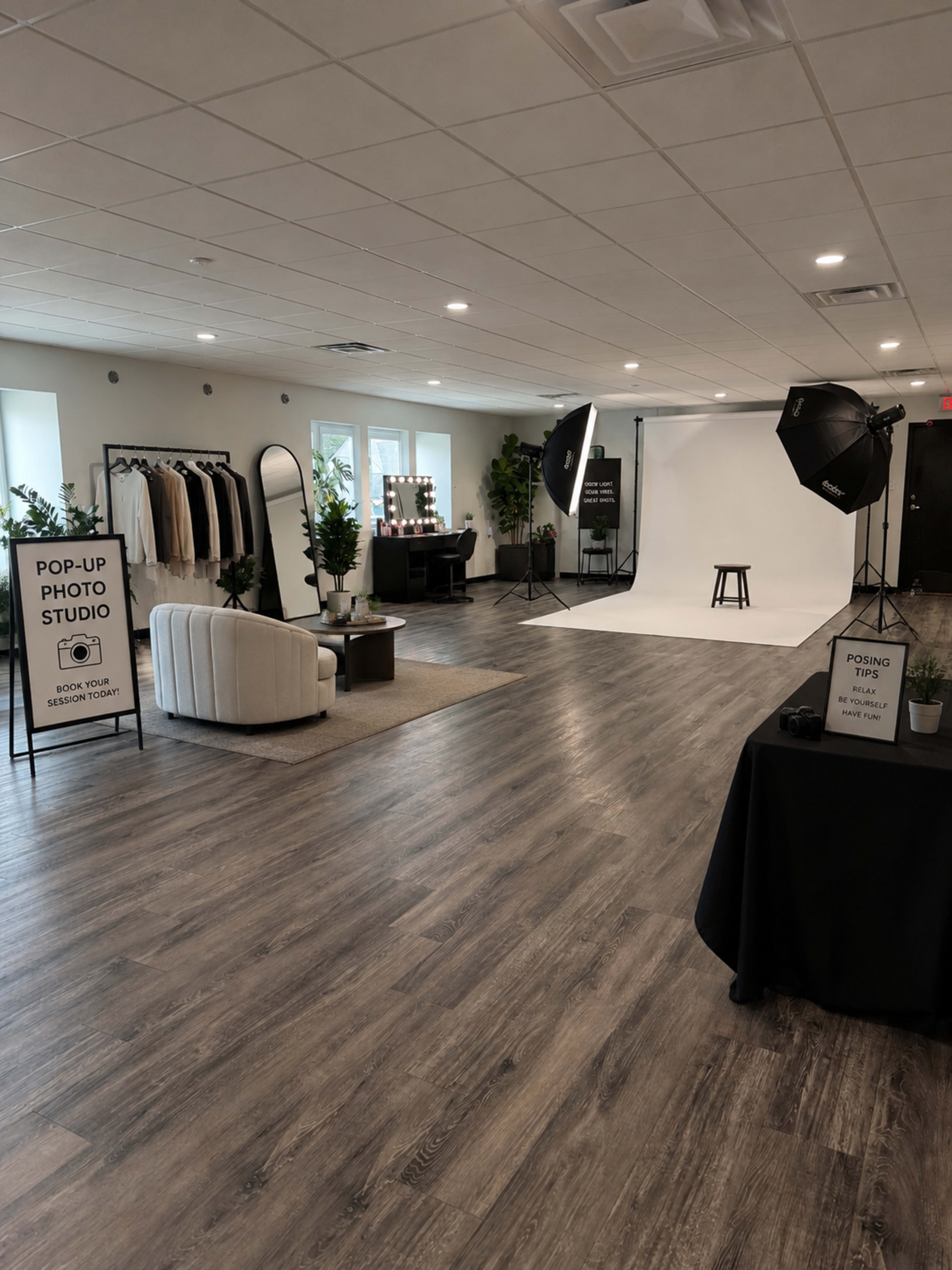 The image shows a spacious pop-up photo studio featuring a white backdrop, studio lights, a stool, and neatly arranged clothing displays along with seating and signage.