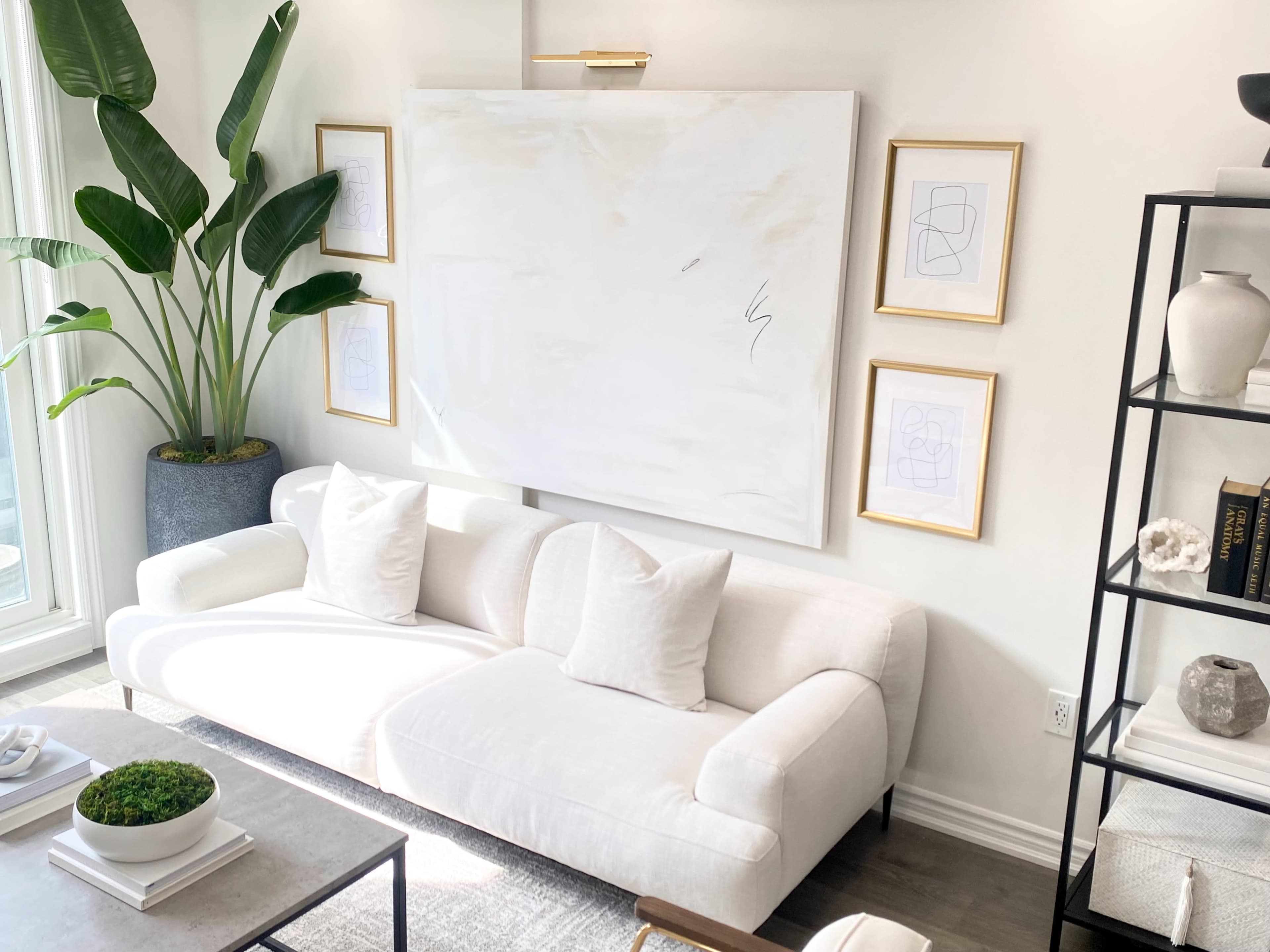 A modern living room features a white sofa with two cushions, a large abstract artwork on the wall, and decorative plants beside framed line drawings.