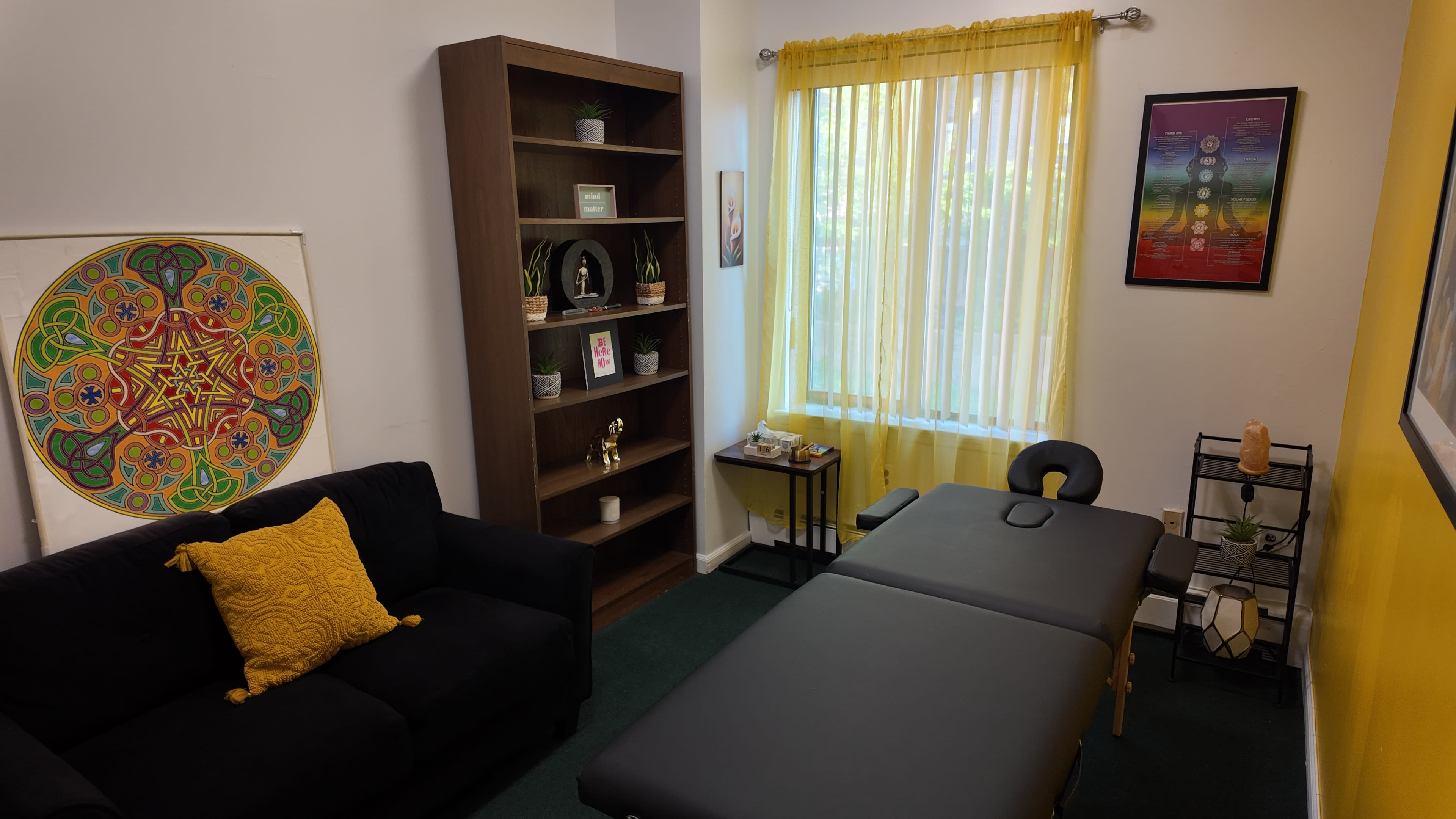 The image shows a therapy room featuring a massage table, a dark couch with a yellow pillow, a bookcase filled with plants and decor, and a large window with yellow curtains.