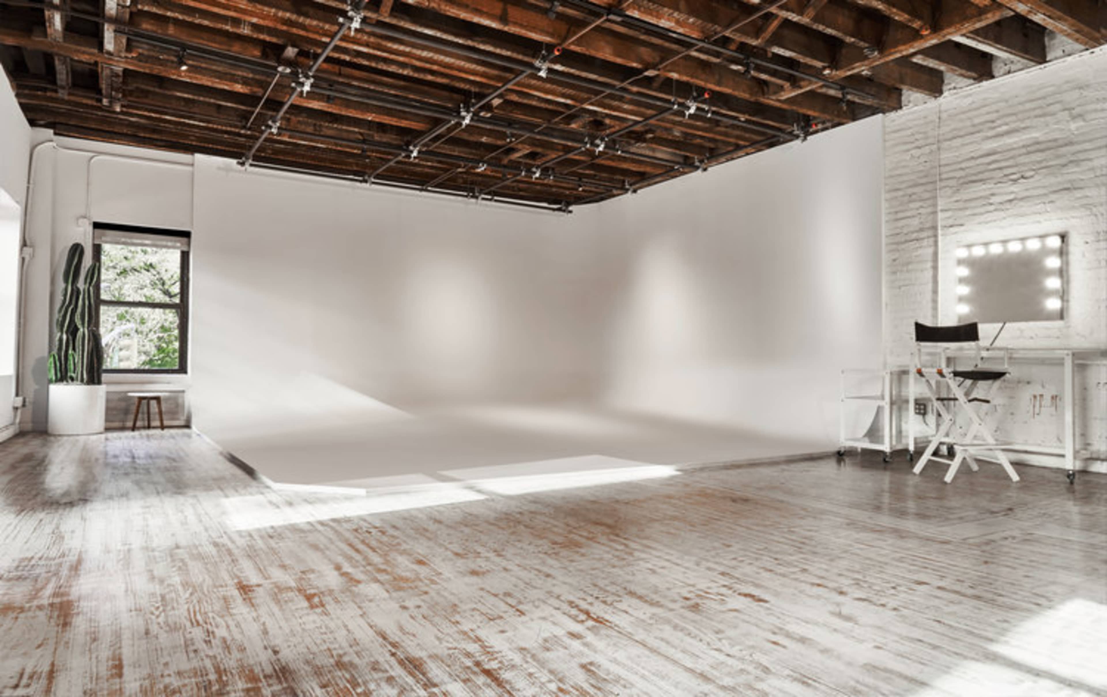 A spacious, well-lit studio with a white backdrop, hardwood floors, and a vanity mirror with lights.