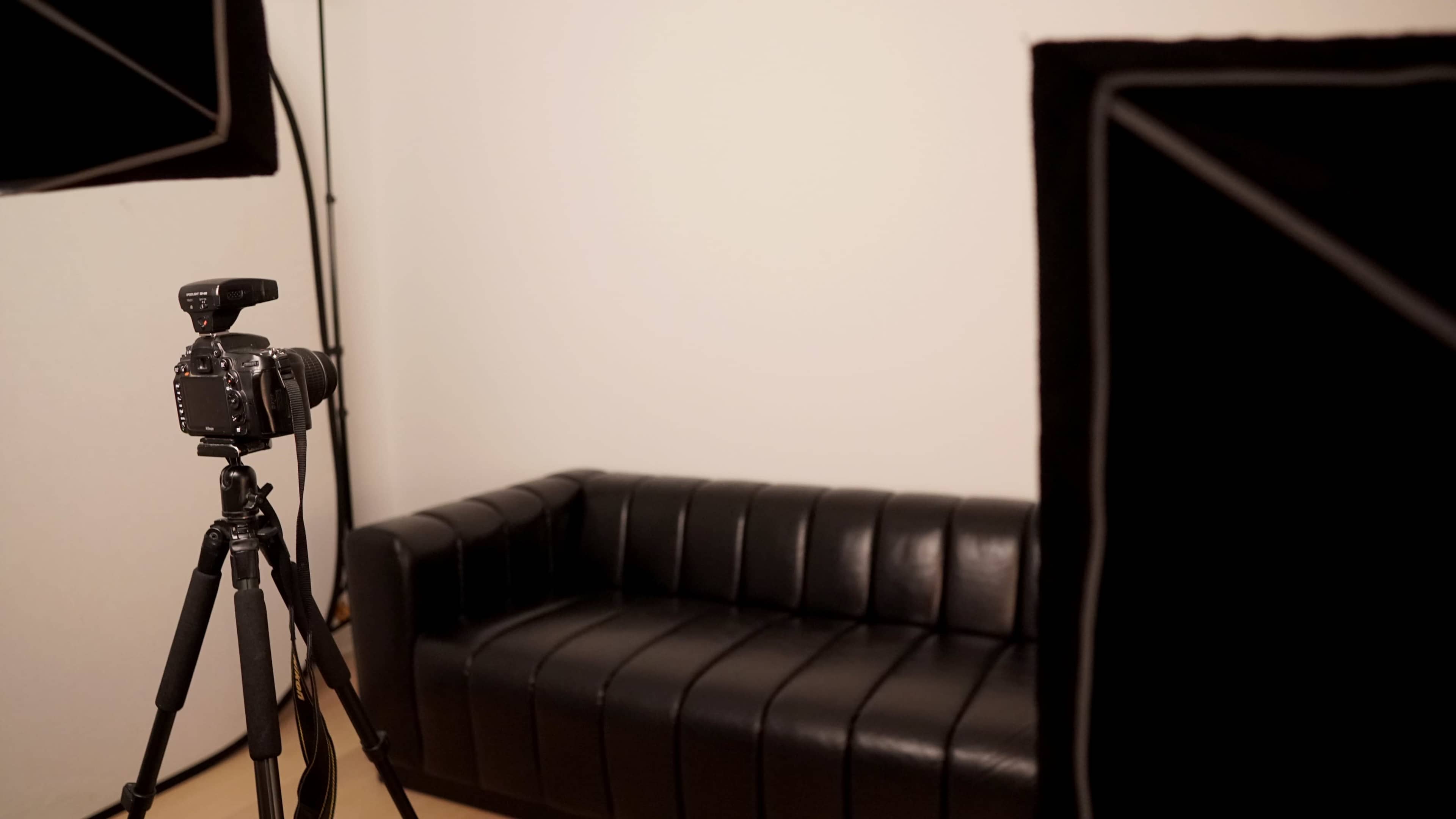 A black leather couch is positioned in a minimalistic studio setting, with a camera on a tripod and lighting equipment nearby.