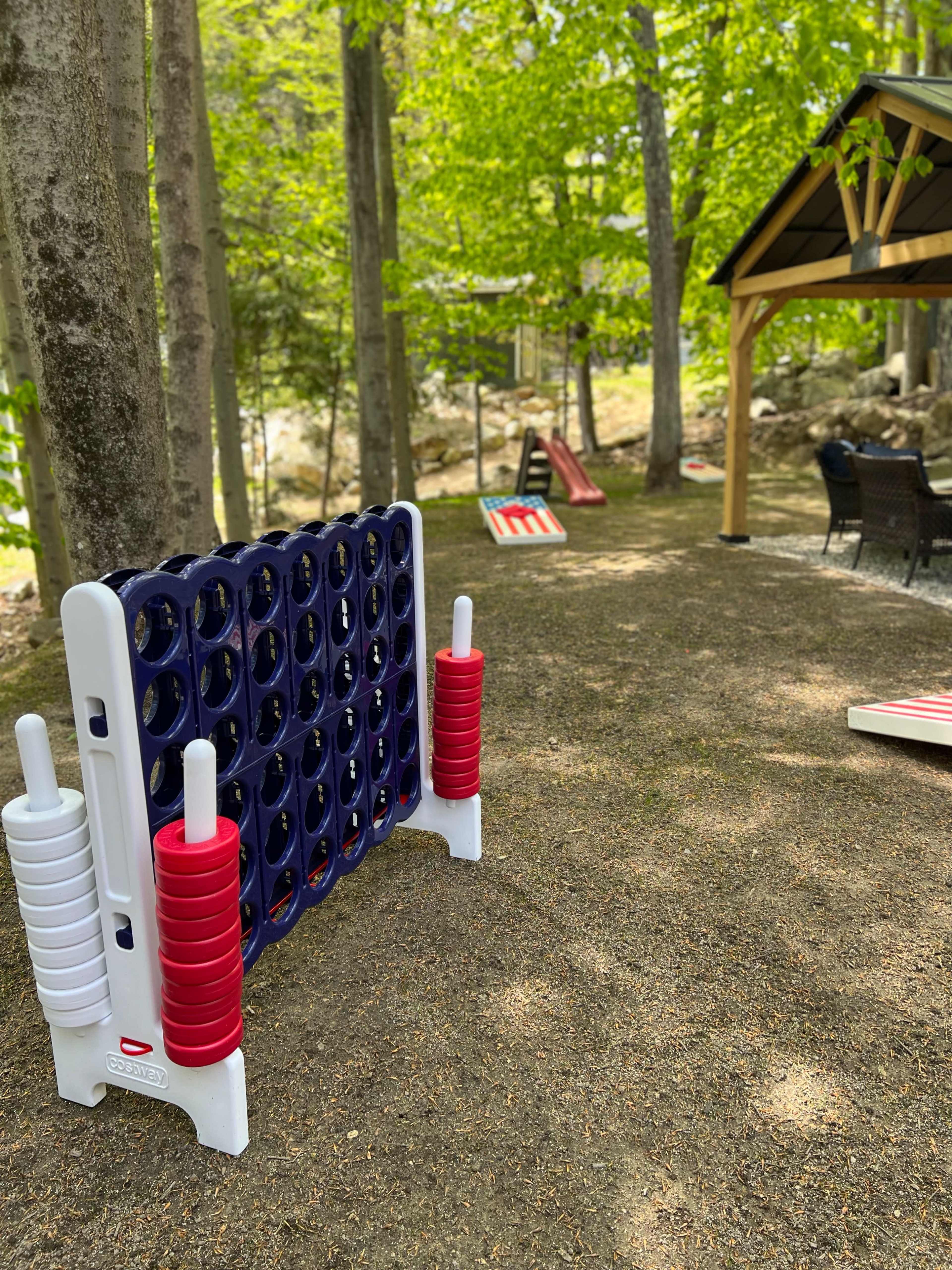 A backyard recreational area featuring a large Connect Four game, a sloped slide, and a seating area under a gazebo surrounded by trees.