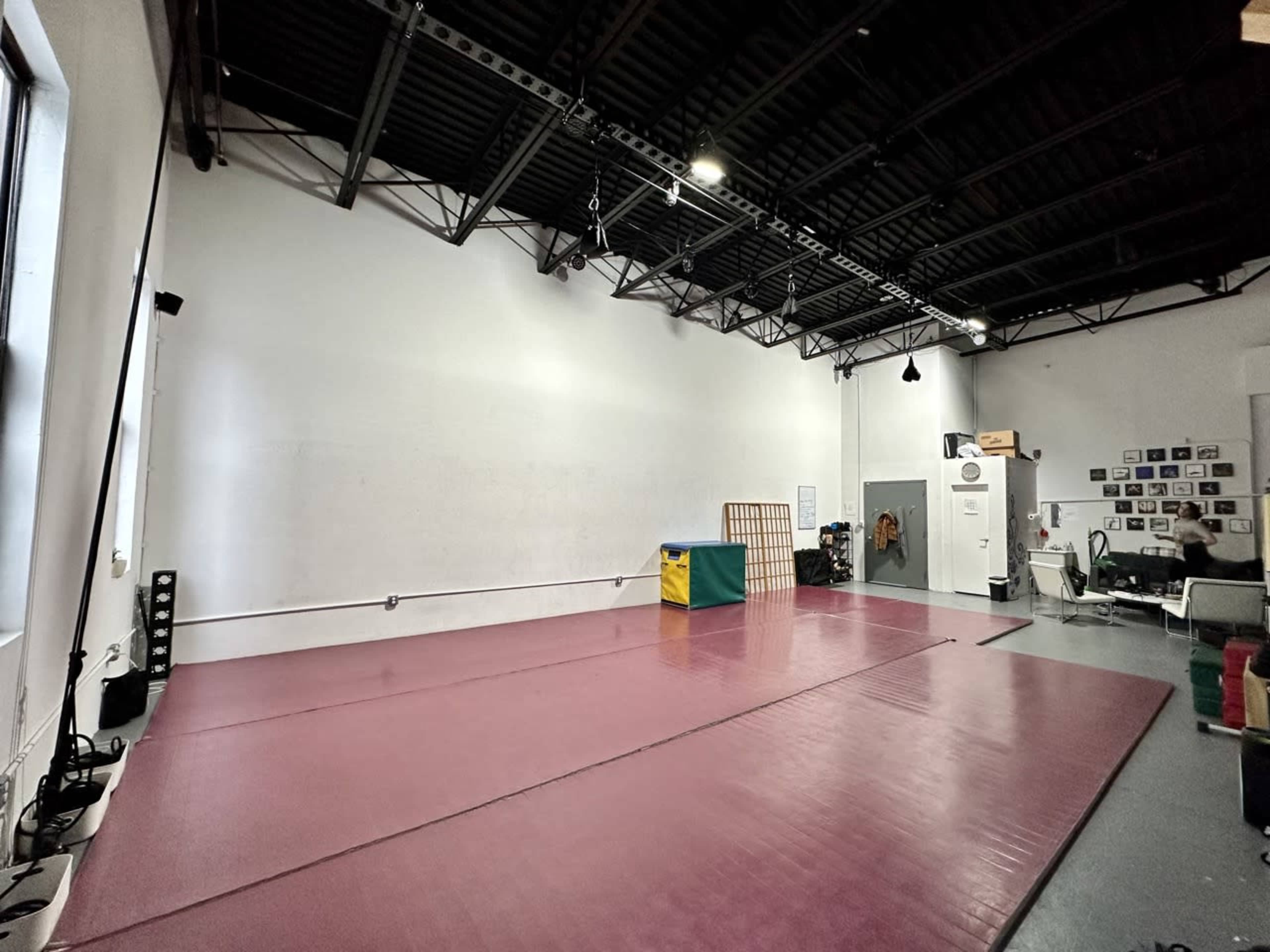 A spacious, empty gymnasium with a pink mat, yellow and green training equipment, and white walls, illuminated by natural light from large windows.