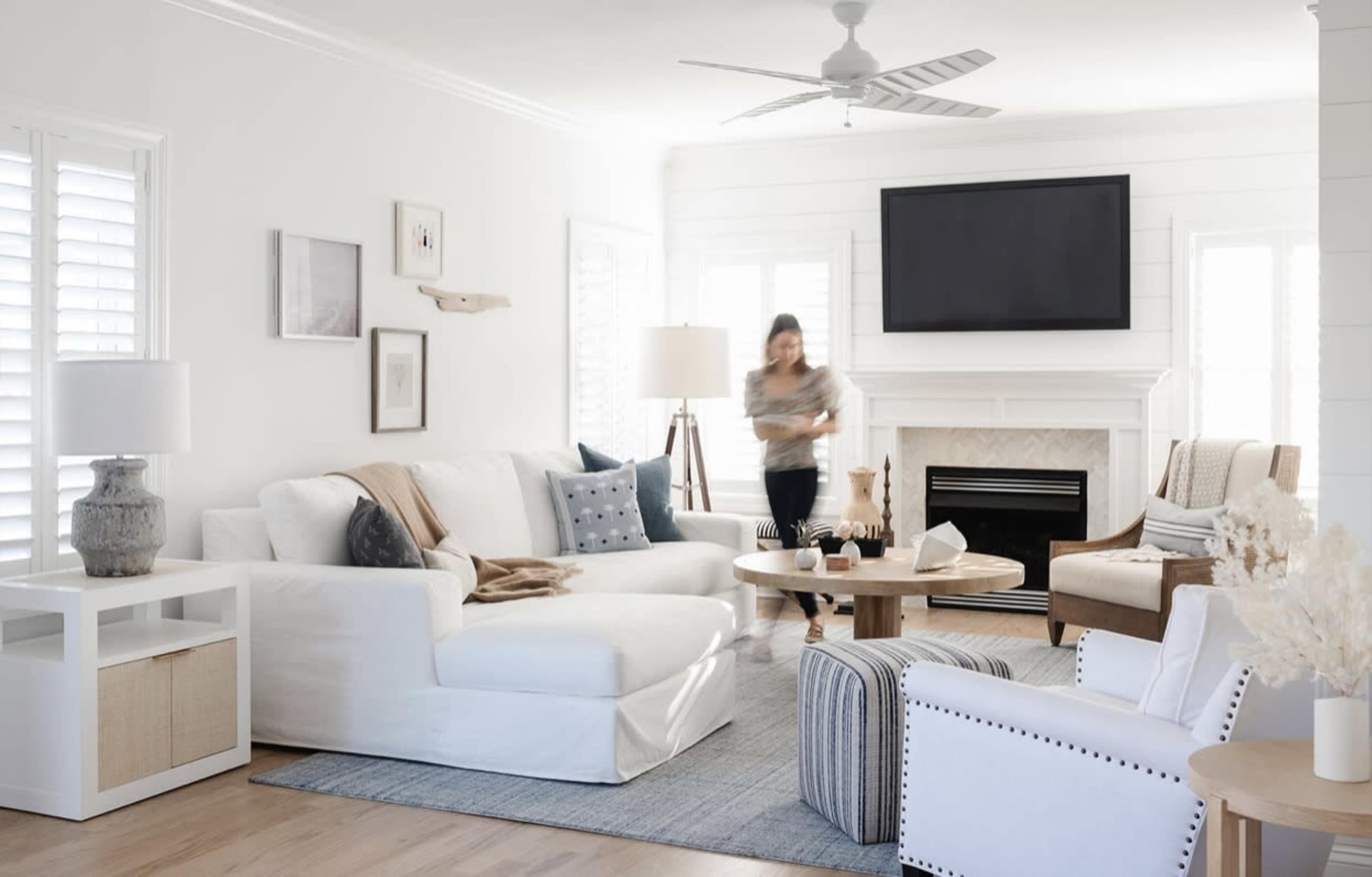 A woman moves through a bright living room featuring a large white sofa, a round coffee table, and a fireplace with a television above it, all surrounded by light-colored decor.