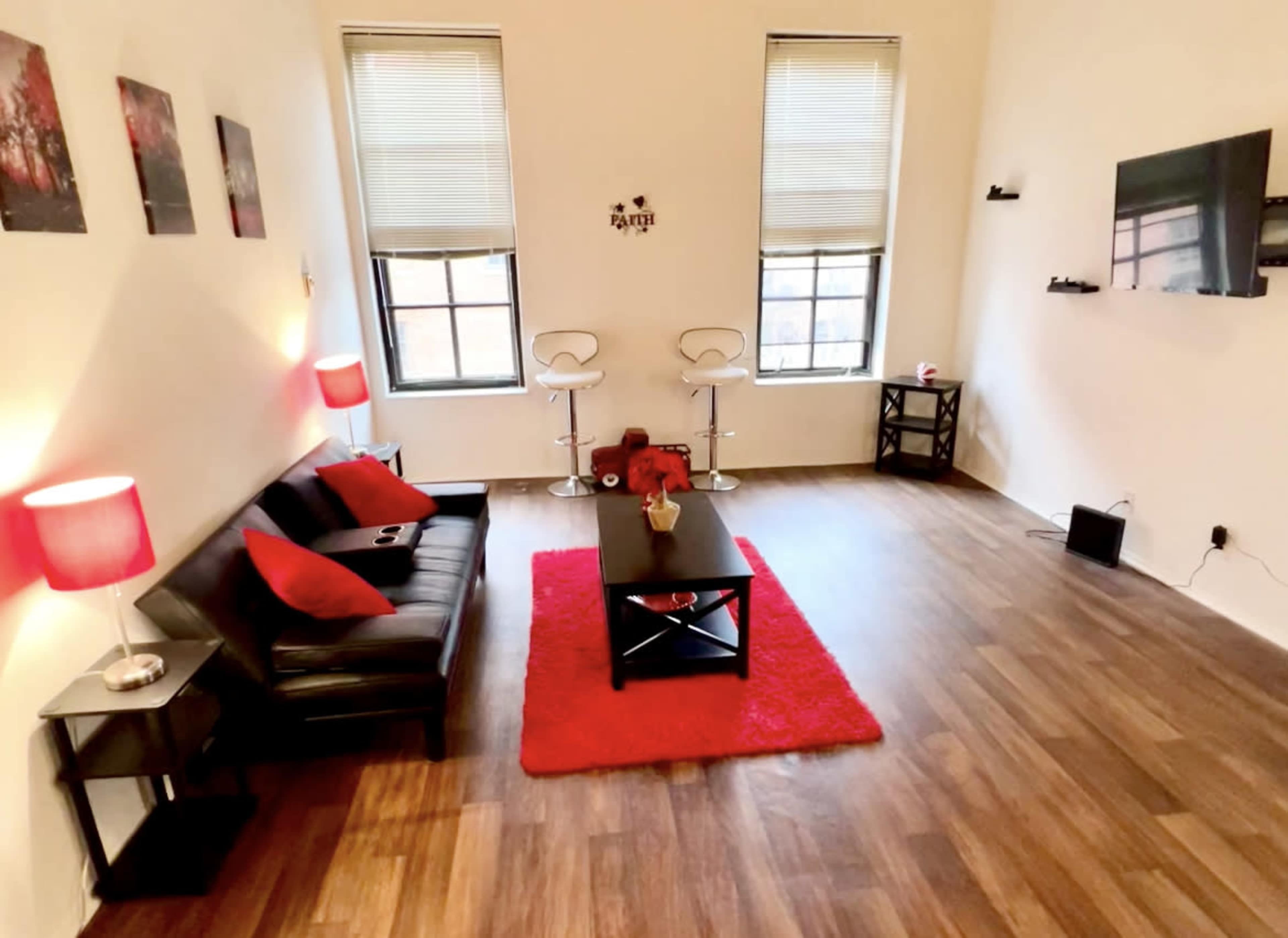 A minimalist living room with a black sofa, a red rug, two bar stools, and a coffee table, featuring large windows with blinds.