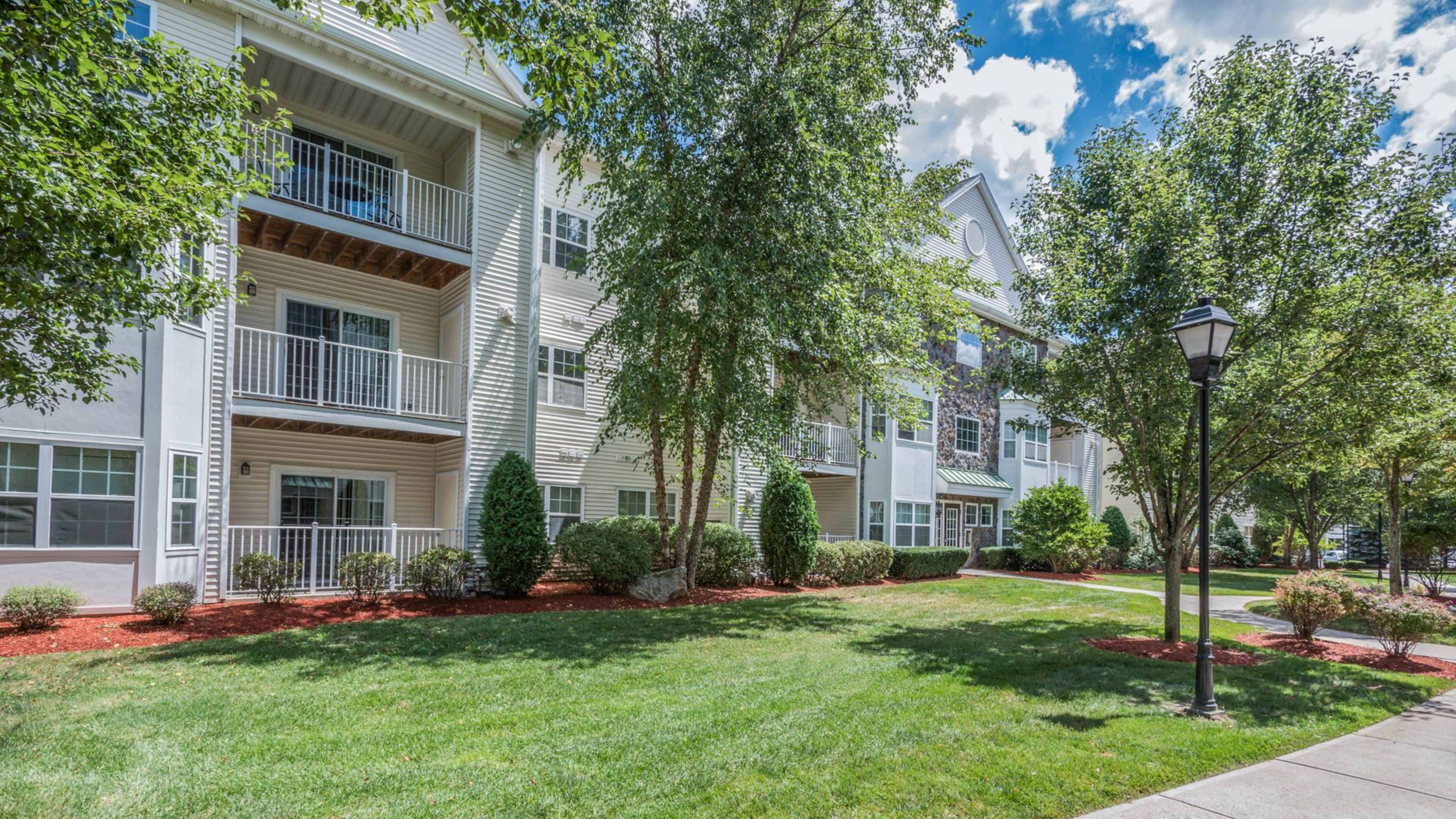 A residential apartment complex with landscaped greenery, trees, and a walkway lined with lamp posts.