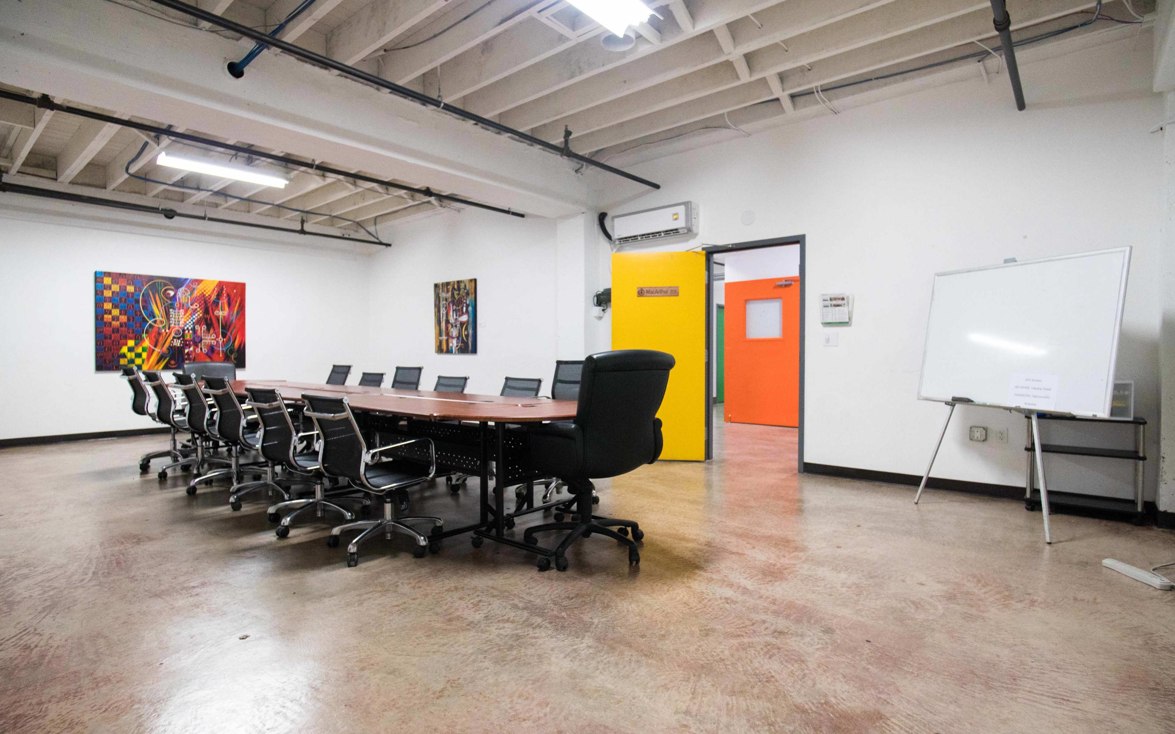 A spacious conference room features a long table with multiple chairs, an easel, and brightly colored artwork on the walls.