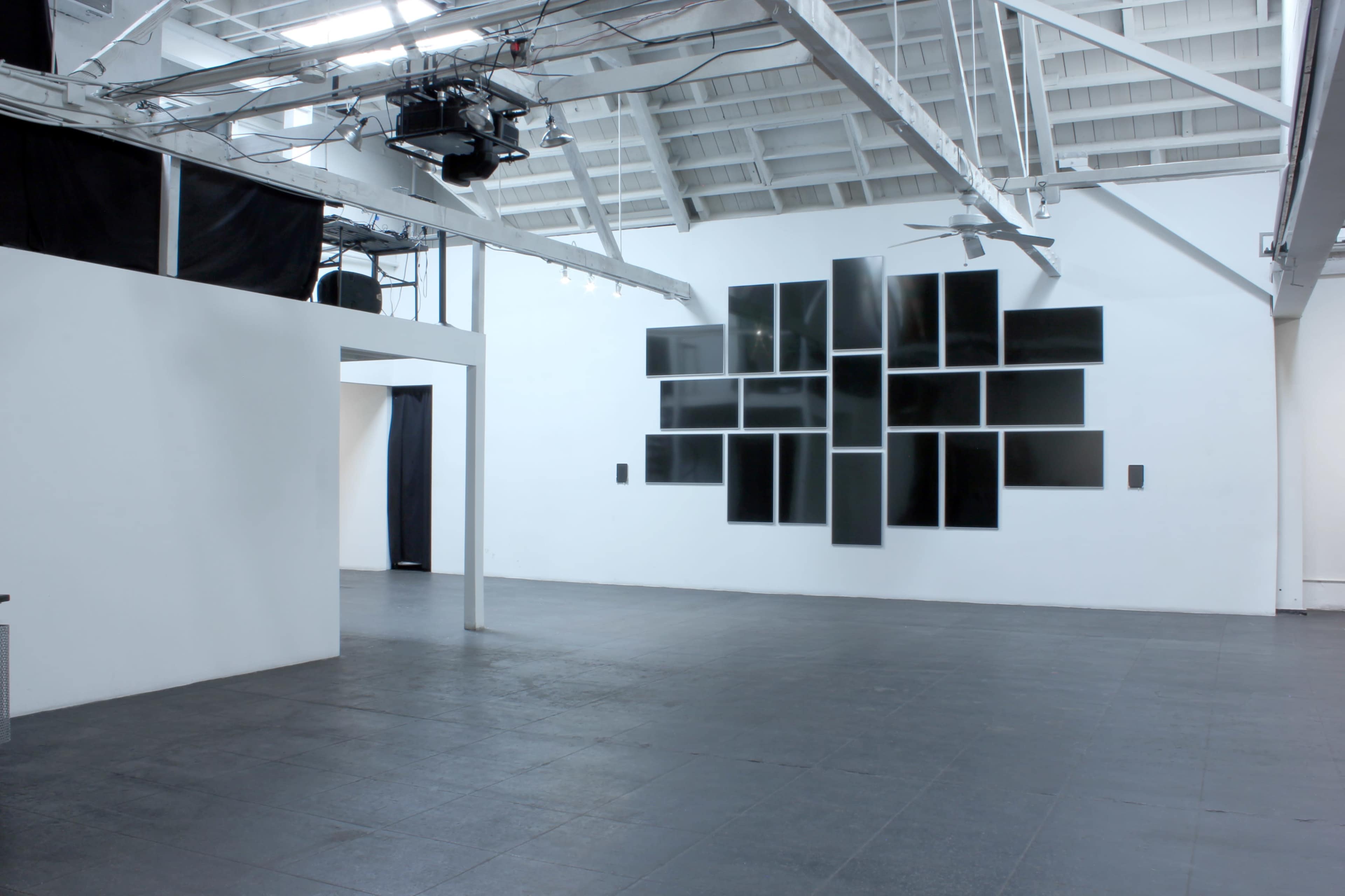 An empty art gallery with white walls, black flooring, and a large arrangement of black panels on one wall.