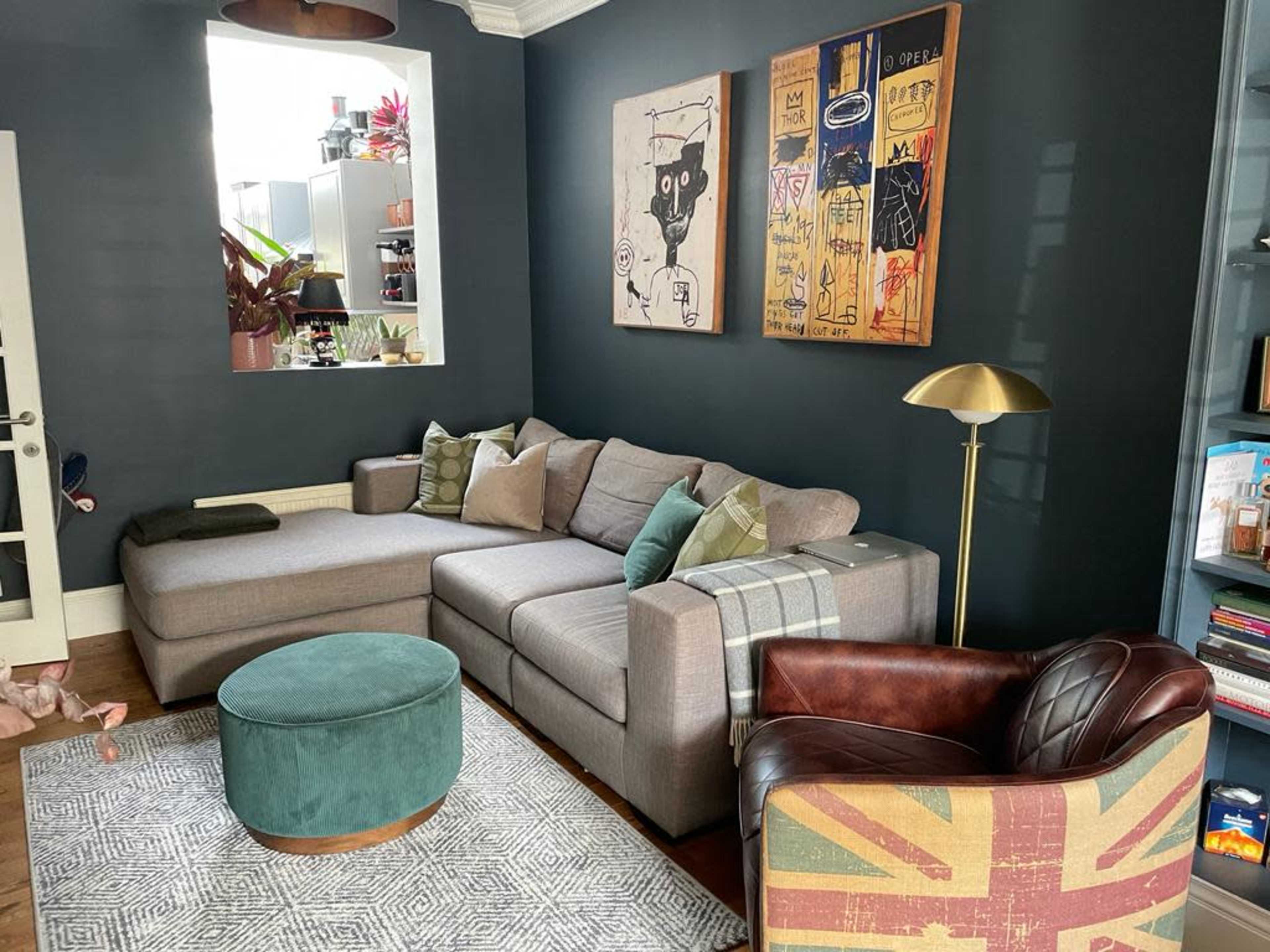 A living room with a gray sectional sofa, a round teal ottoman, and a brown leather chair, complemented by green accents and colorful artwork on the walls.