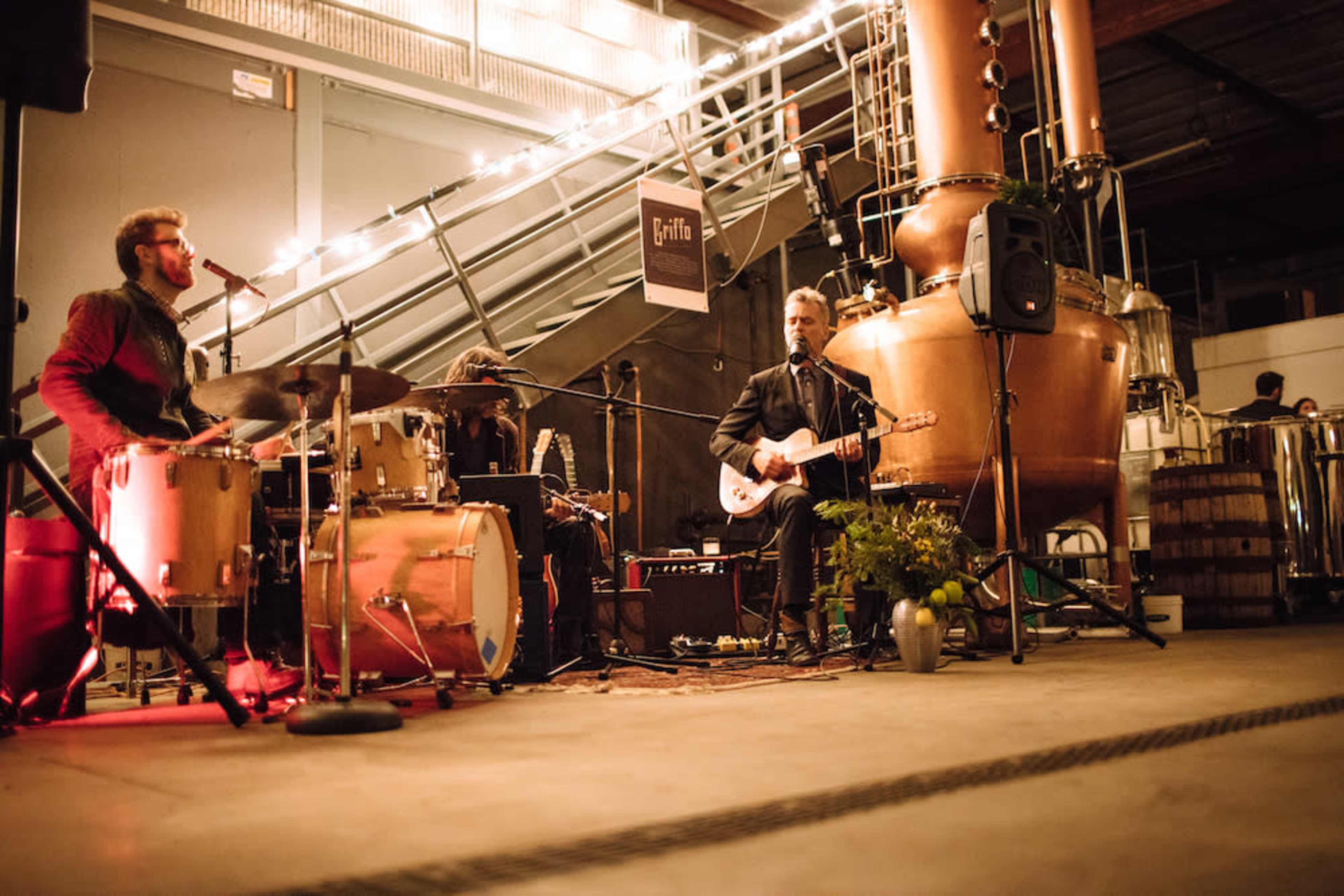 A band performs on a stage set up in a brewery, surrounded by brewing equipment and string lights.