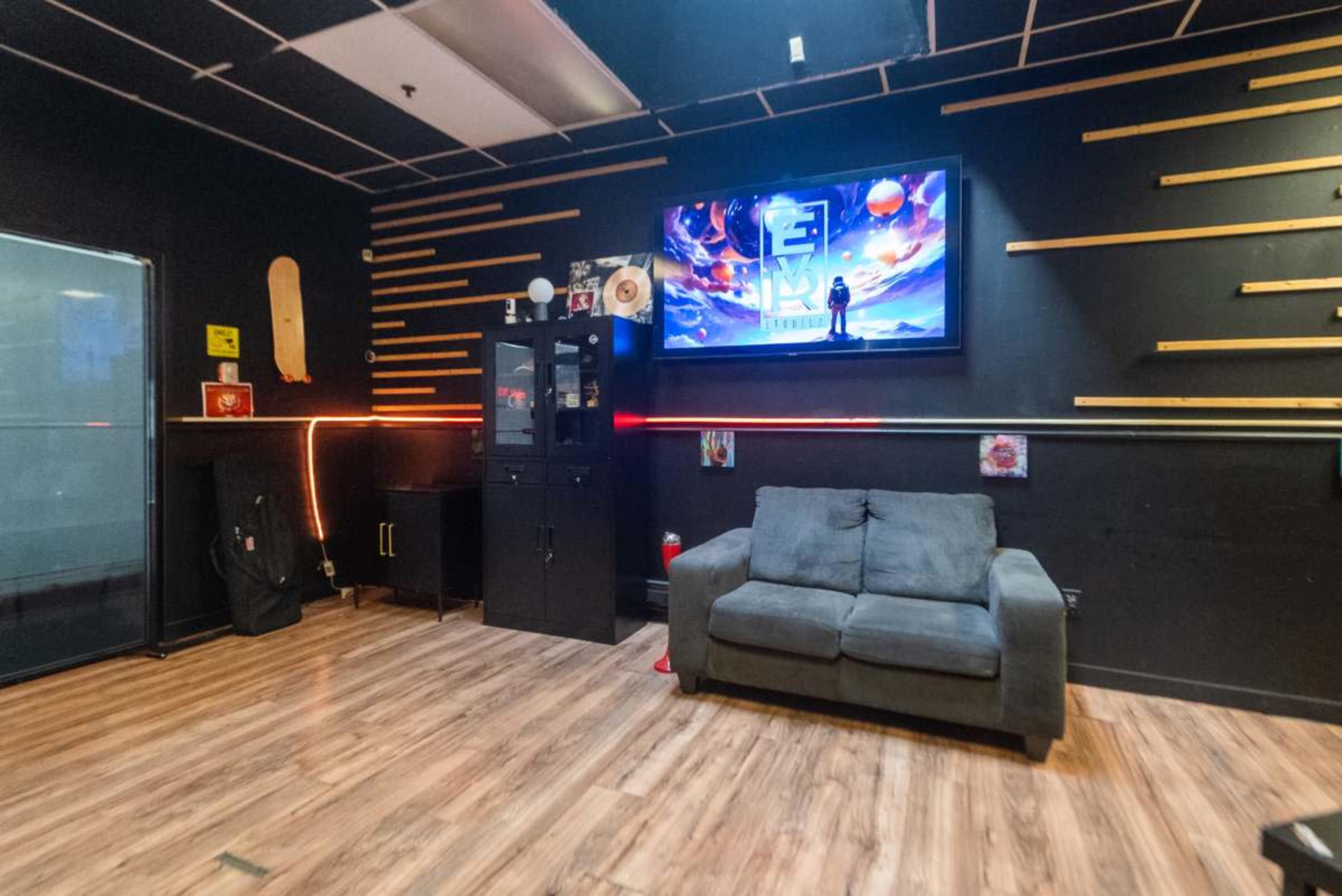 The image shows a dimly lit room with a gray couch, a television mounted on a black wall, and a cabinet next to it, along with wooden wall accents and some decorations.