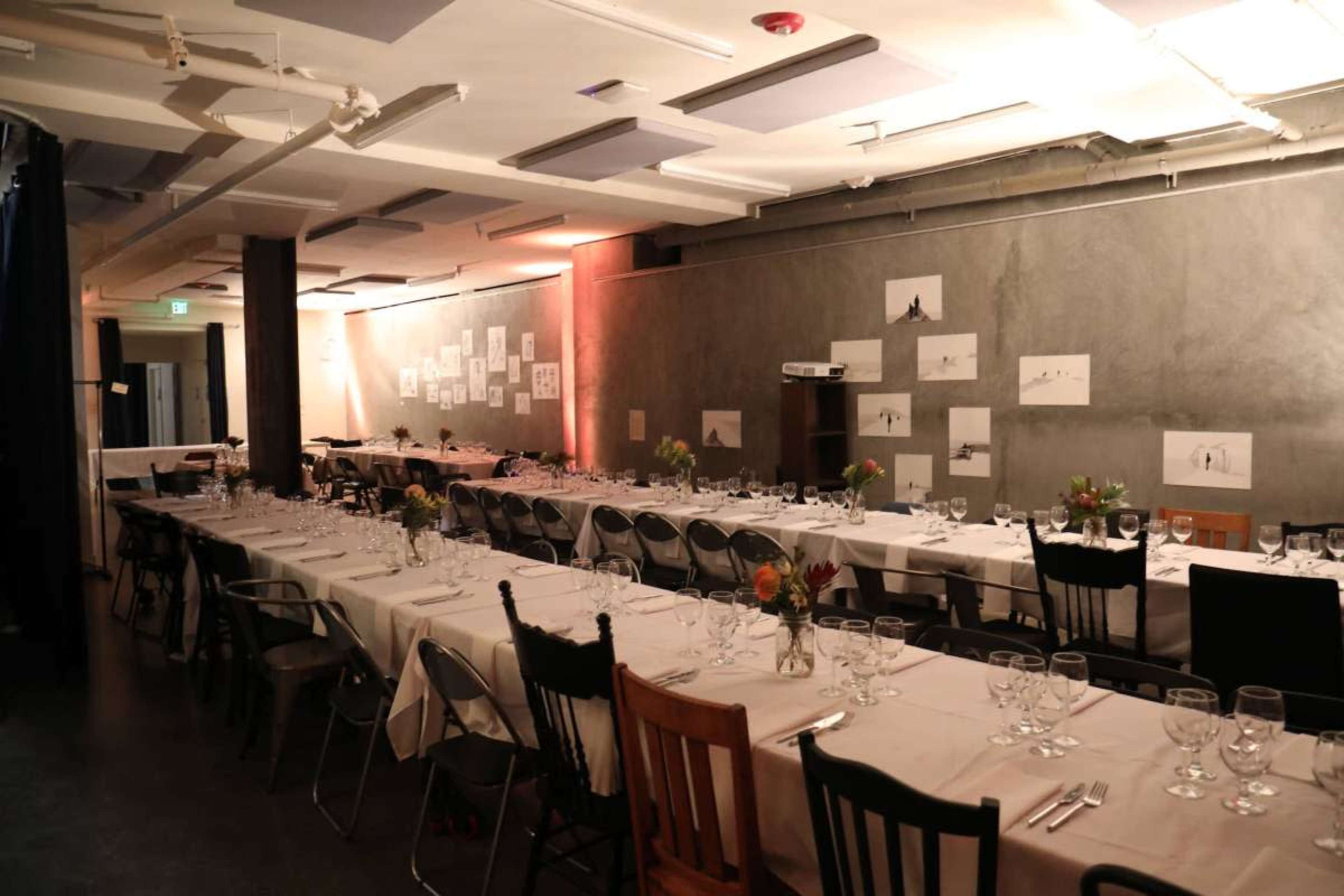 The image shows a dining hall set with long tables covered in white tablecloths, adorned with glassware and floral centerpieces, against a backdrop of minimalist artwork on the walls.