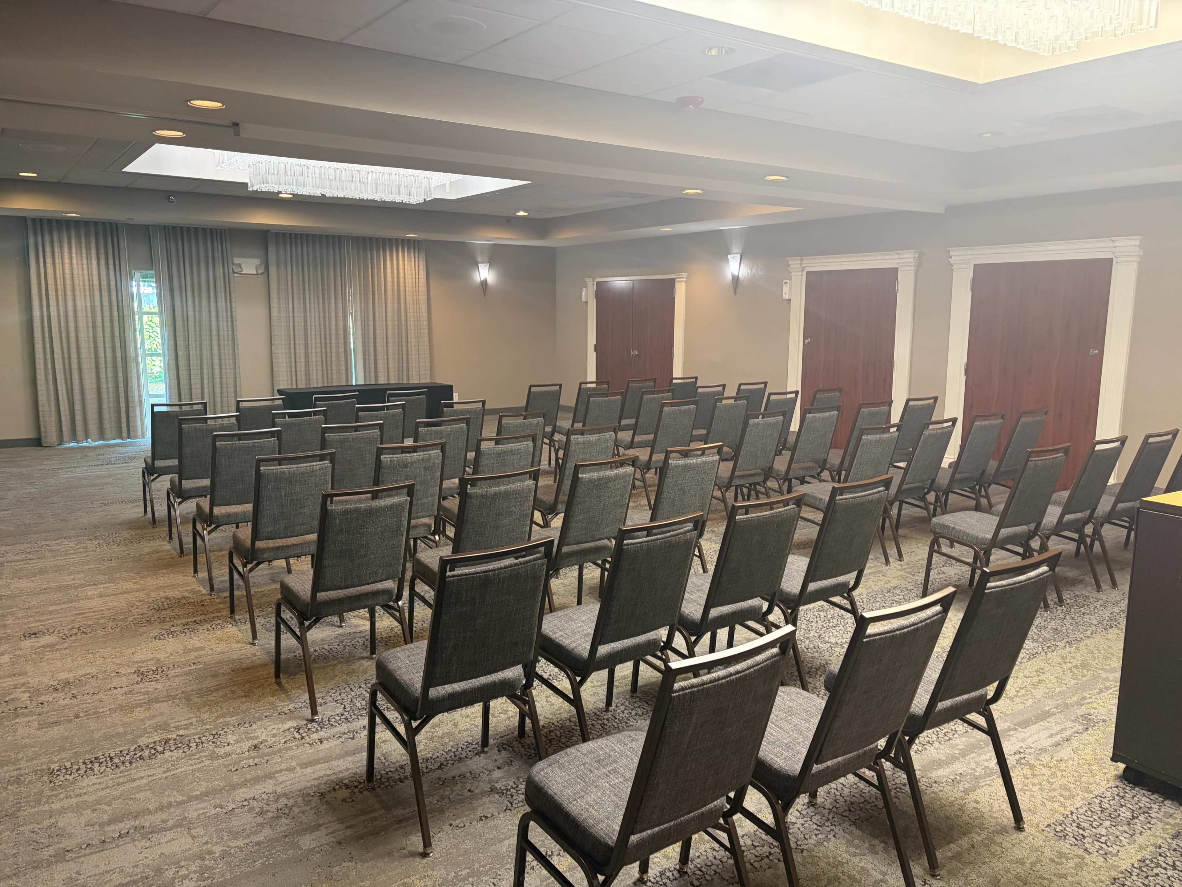 The image shows a conference room set up with rows of gray chairs facing a podium, with doors on either side and windows allowing natural light.