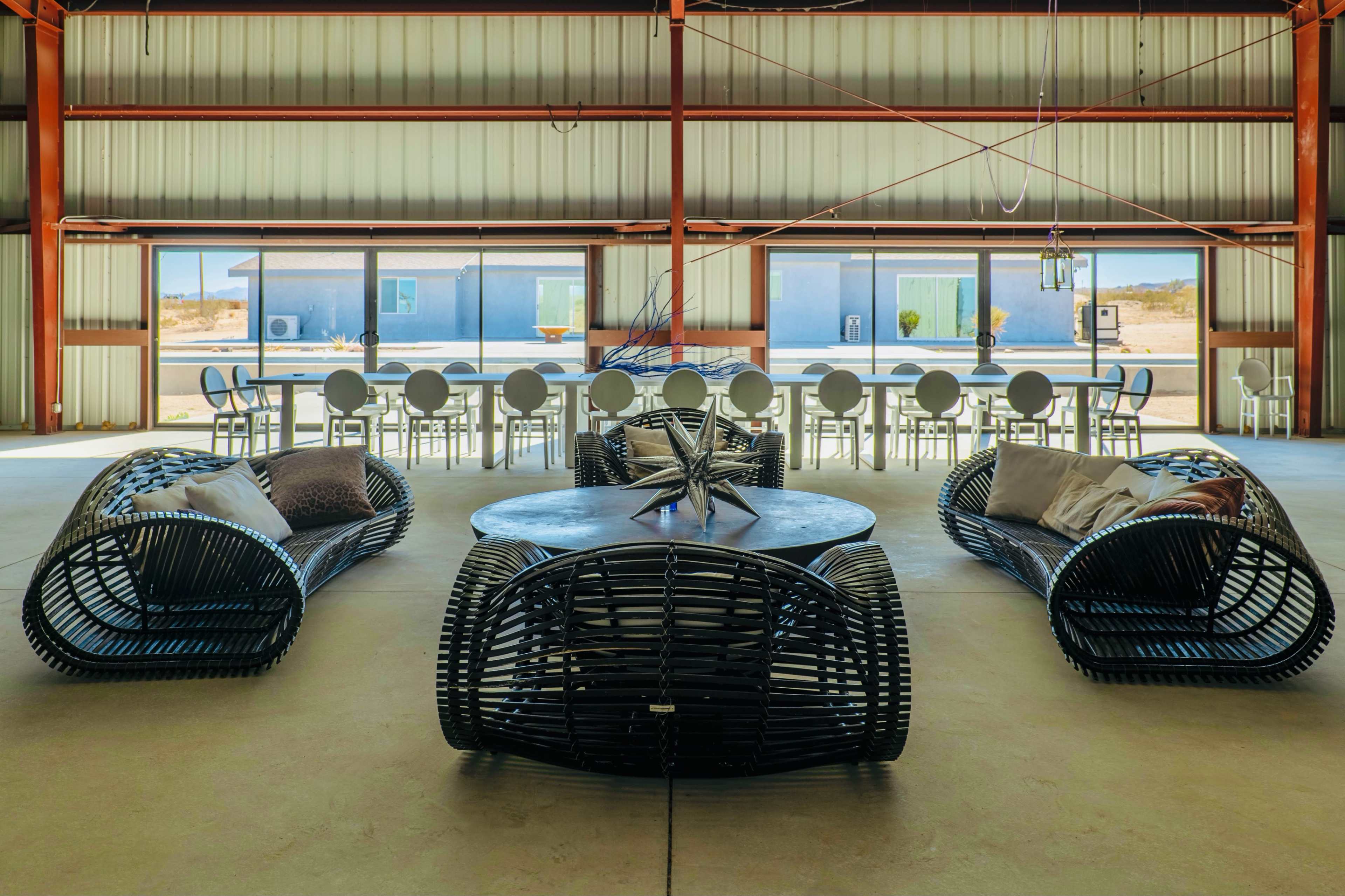 The image shows a spacious indoor area featuring a modern seating arrangement with wicker furniture and a large dining table, surrounded by large windows.