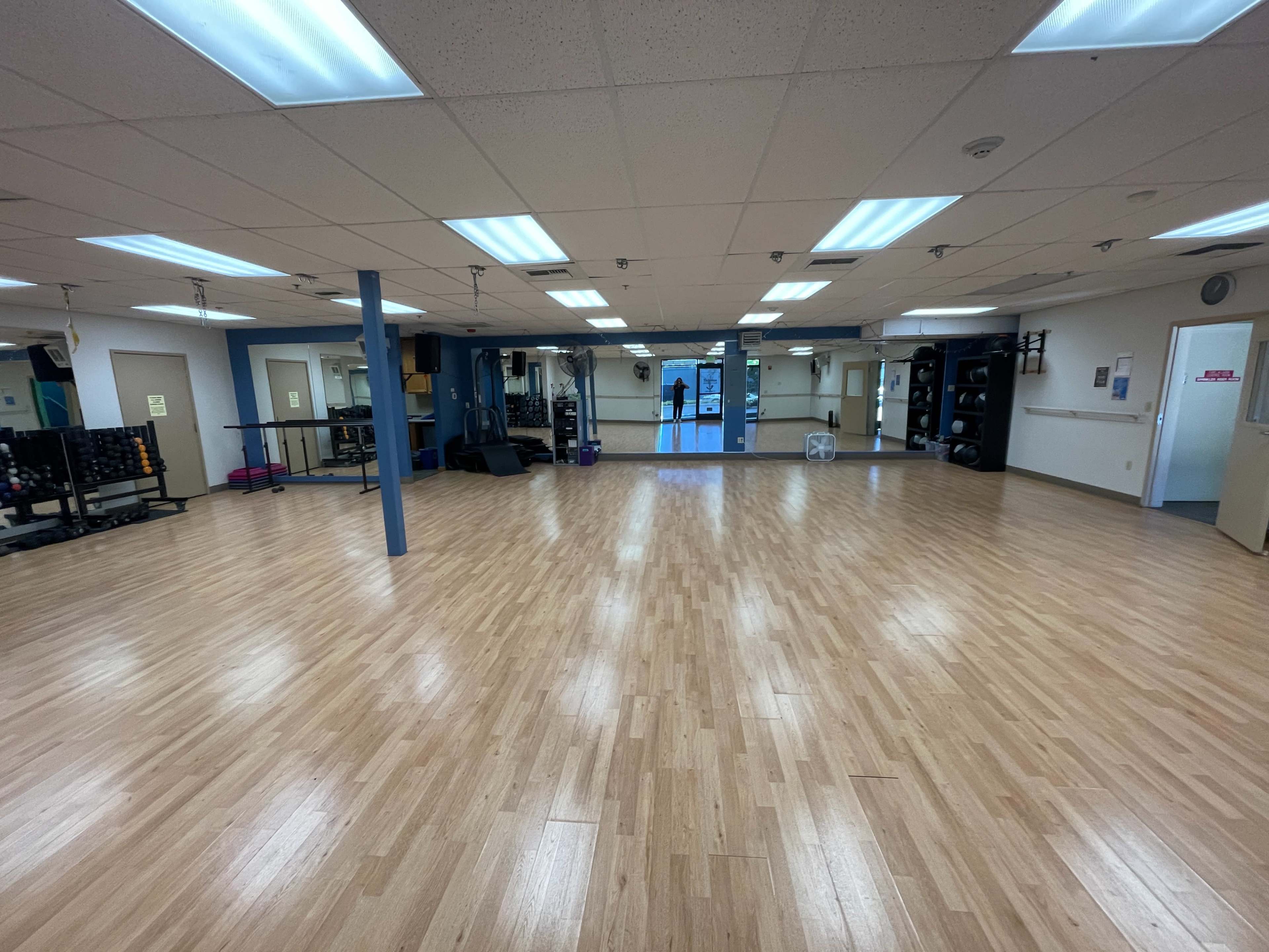 A spacious, clean exercise studio featuring wooden flooring, mirrors, and fitness equipment along the walls.