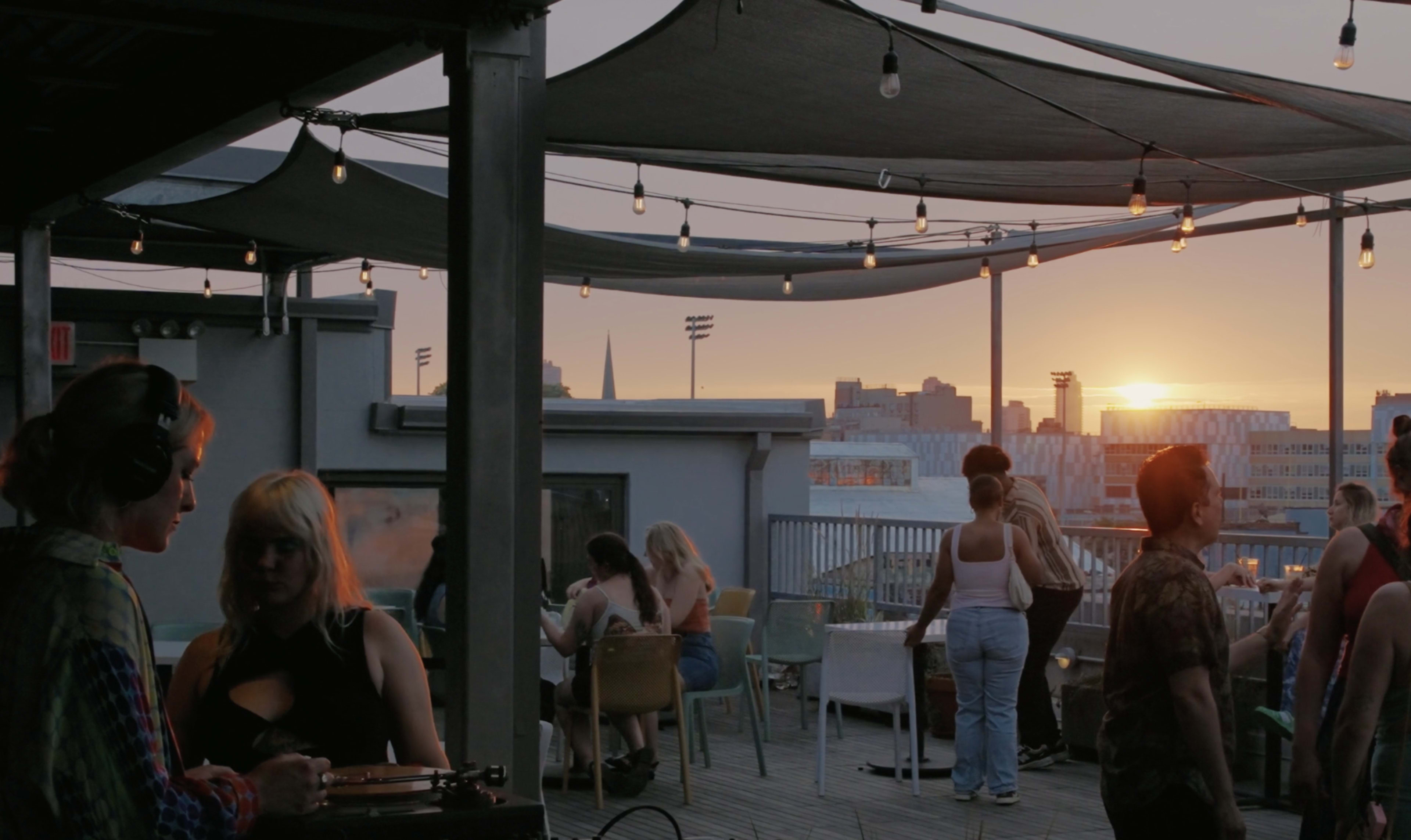 The image shows a rooftop venue with several people socializing and a sunset in the background, with lights strung across the area.