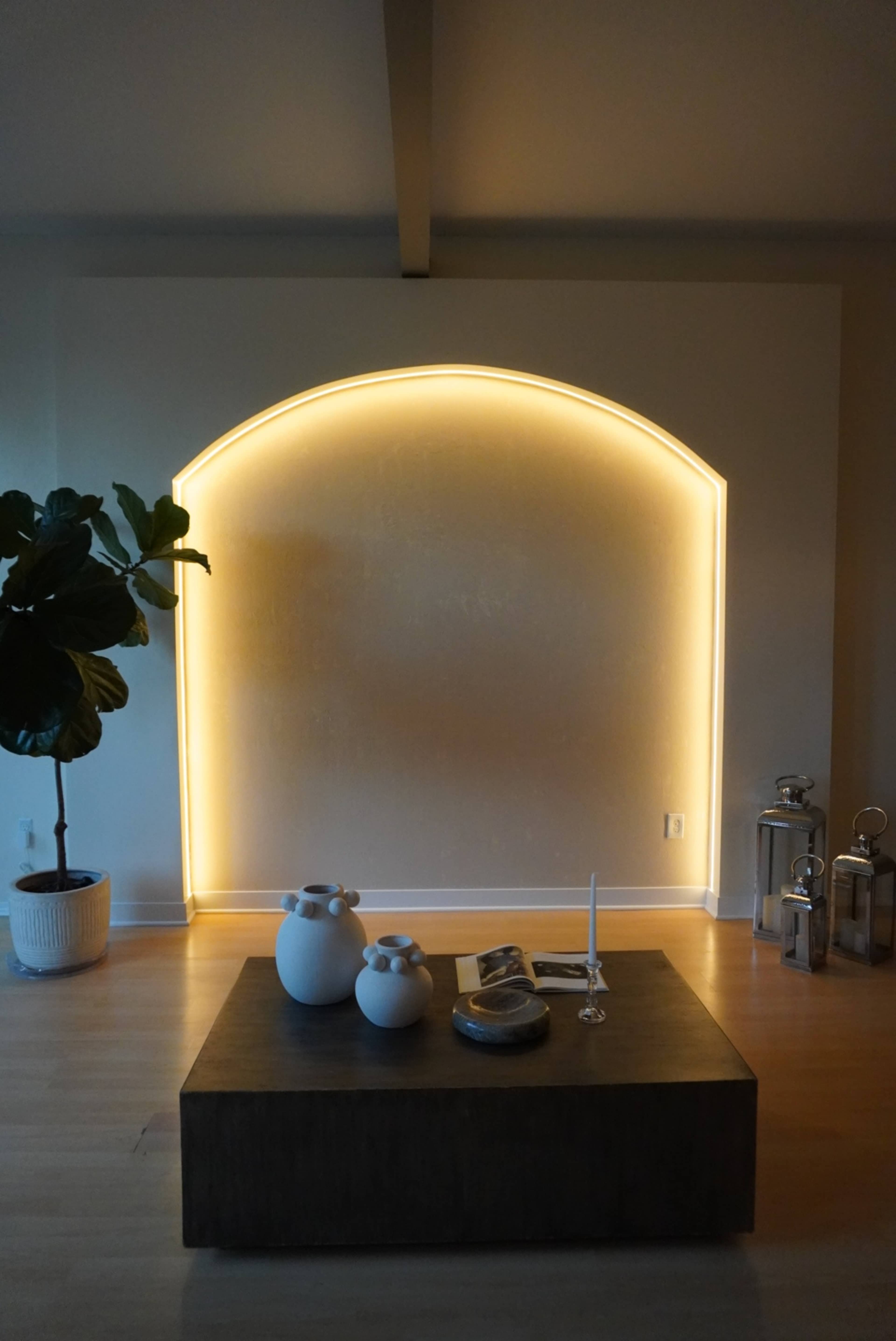 The image features a modern living space with a softly lit arch-shaped wall, a wooden table with decorative items, and a large potted plant in the corner.