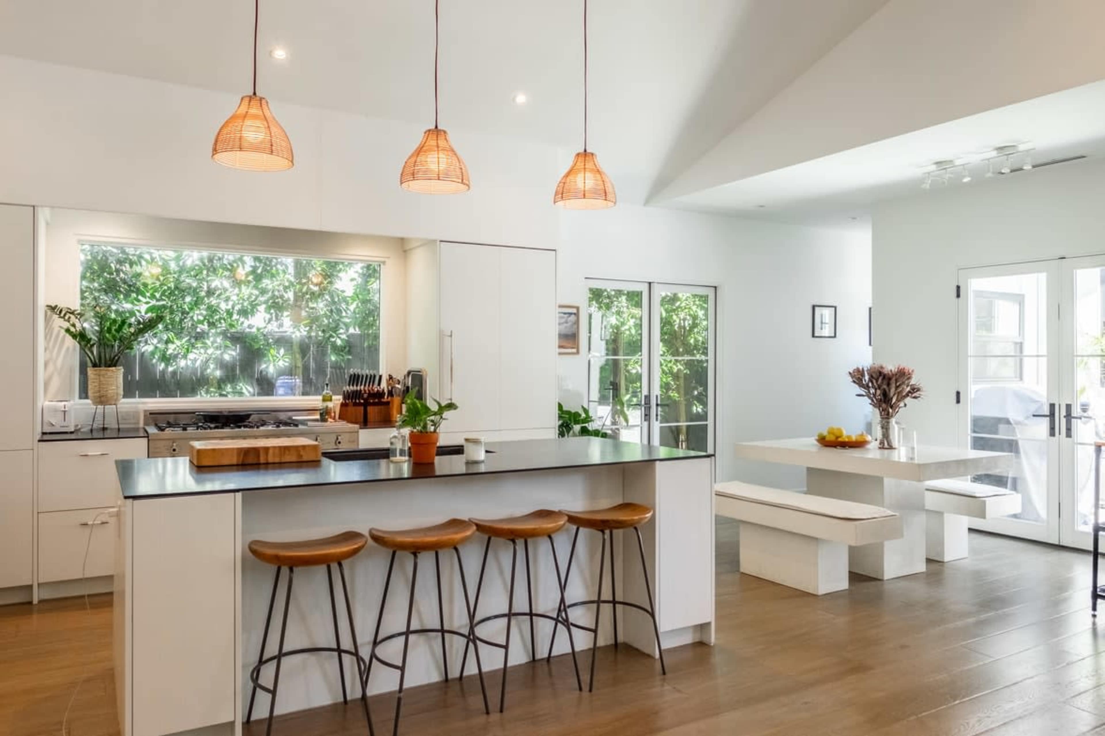A modern kitchen with a large island, three wooden stools, pendant lighting, and an adjacent dining area featuring a long white table.