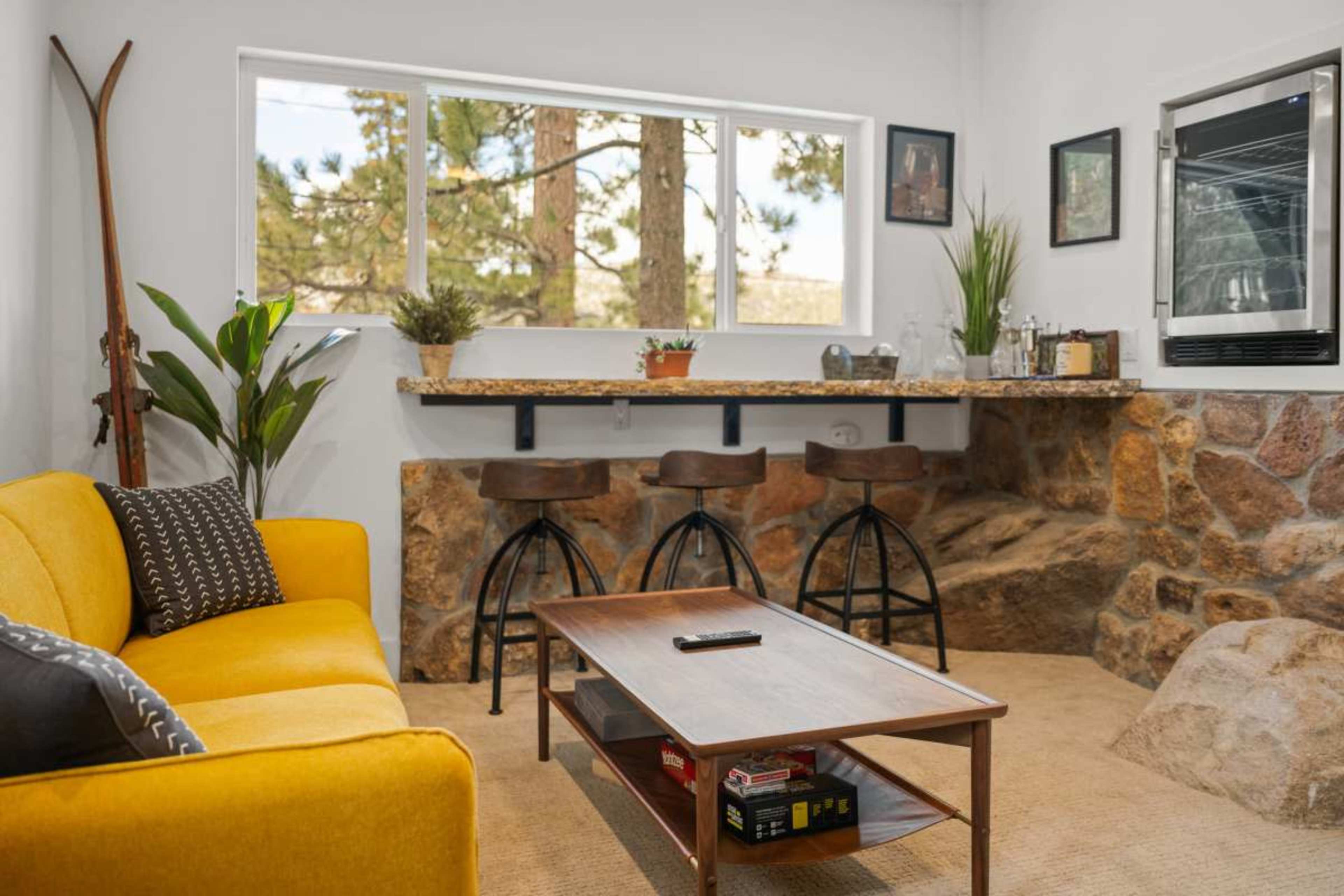 A cozy living space featuring a yellow sofa, a wooden coffee table, and a bar area with stools made of stone and wood, illuminated by natural light from a large window.