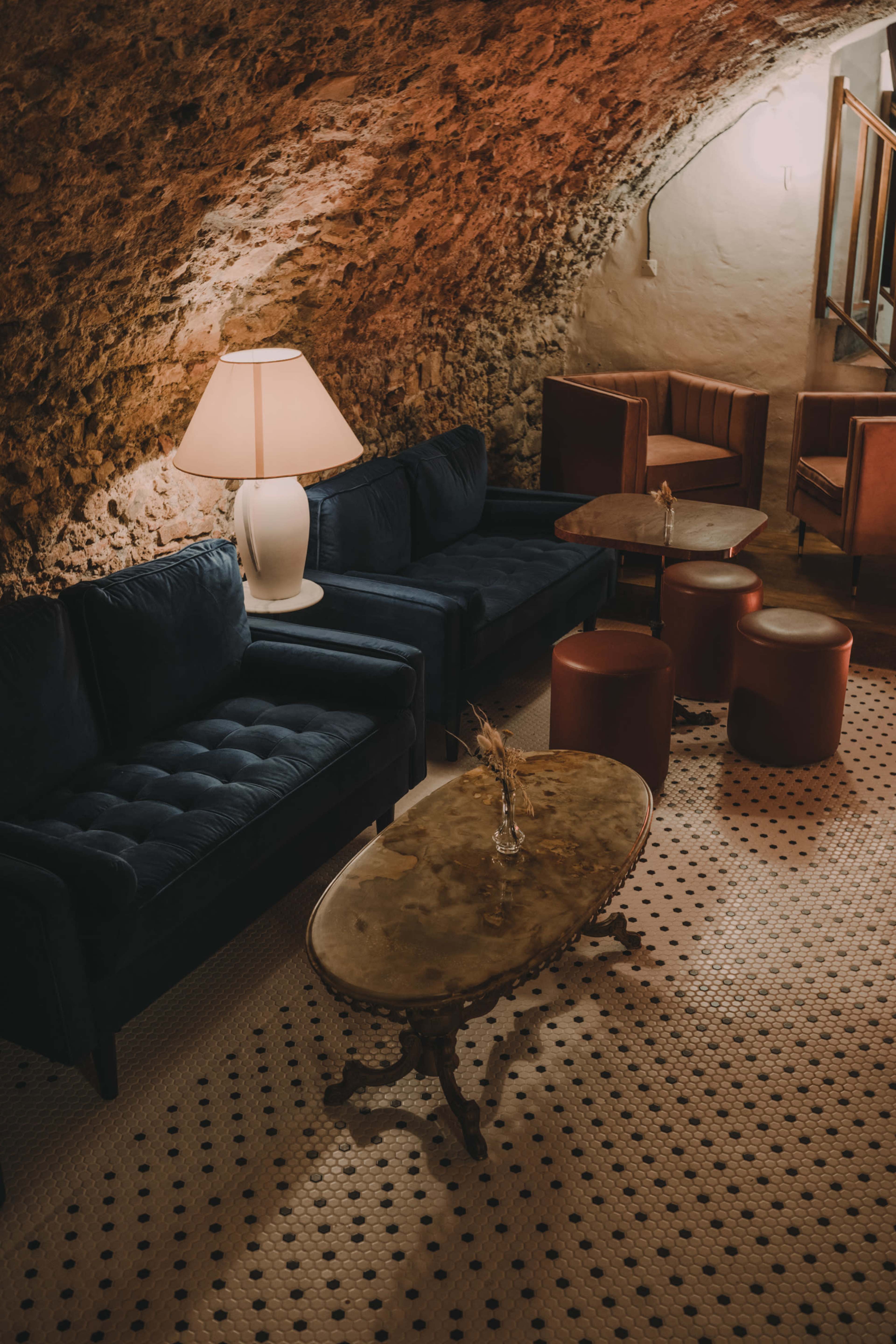 A cozy lounge area features plush blue sofas, elegant wooden tables, and a vintage lamp under a stone archway.