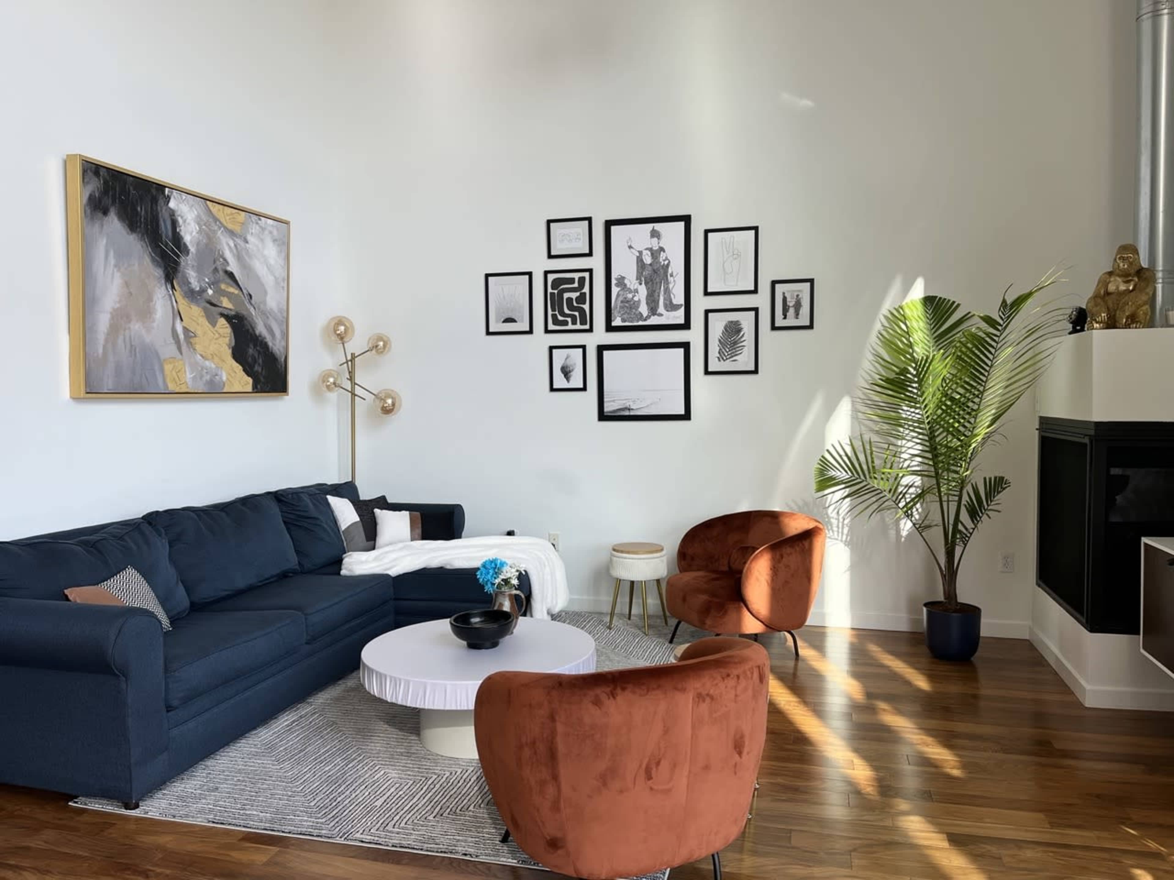 A modern living room features a dark blue sofa, two orange armchairs, a round white coffee table, and a wall adorned with black and white framed artwork, alongside a tall green plant.