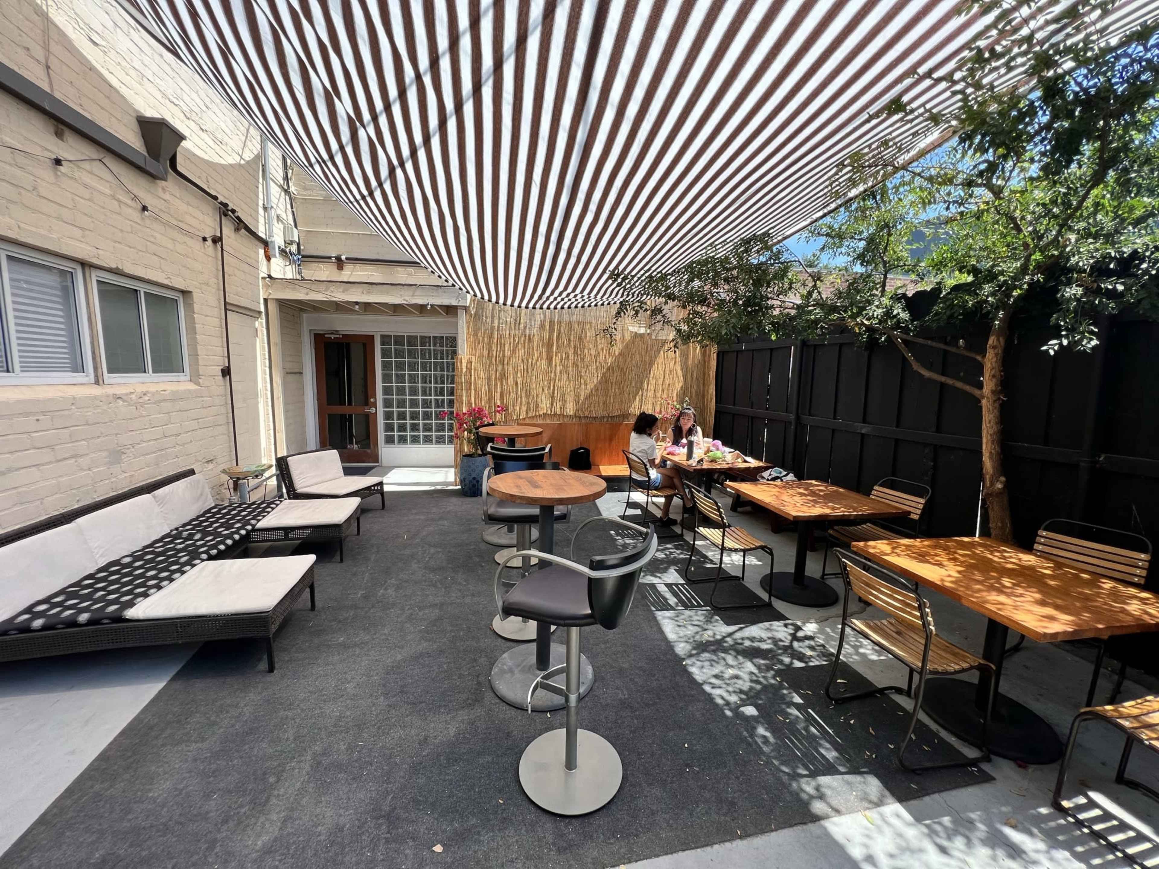 A backyard patio area with striped overhead shading, tables, and seating arrangements, where two people are sitting at a table.