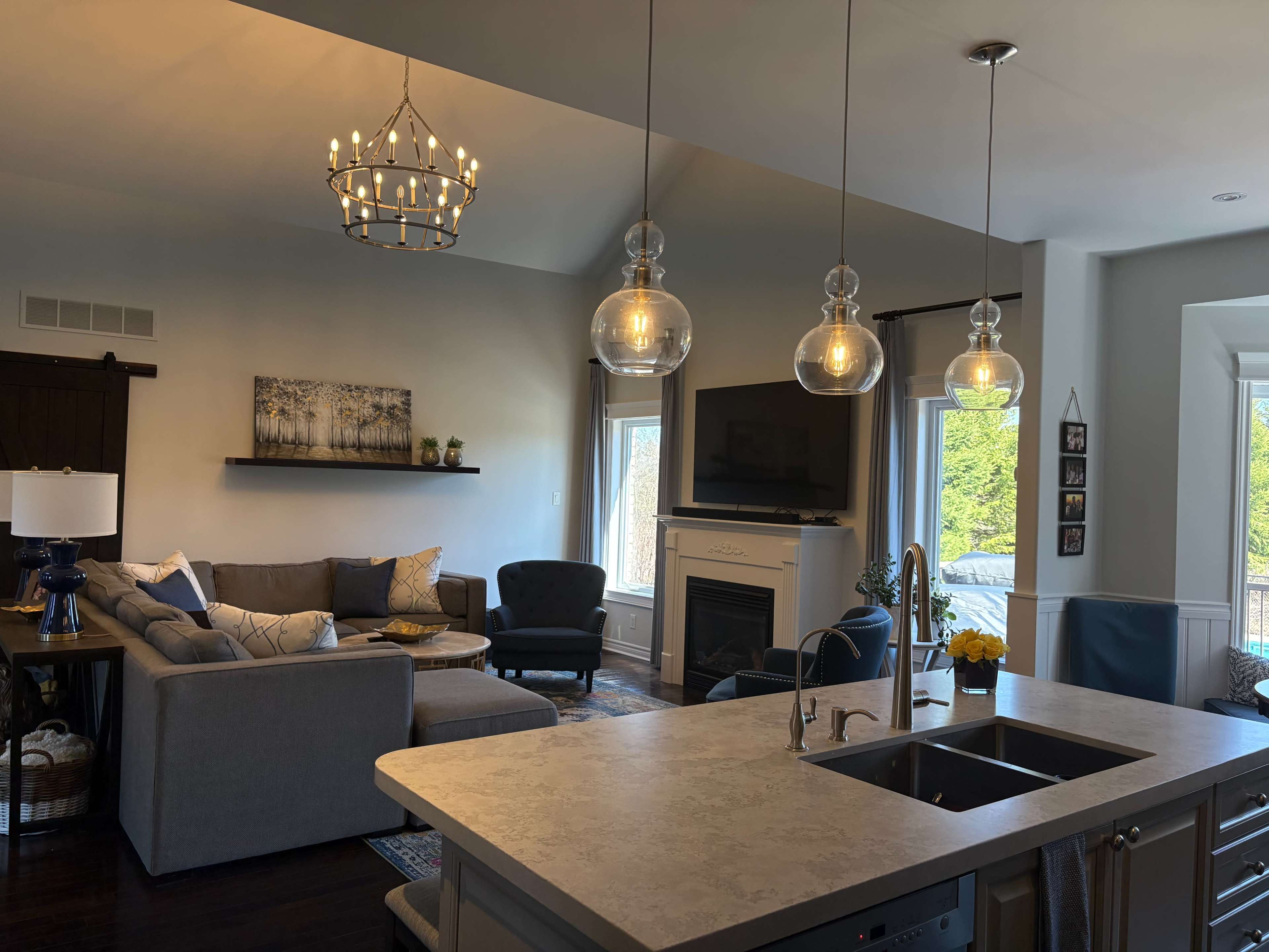 The image shows a modern living room connected to a kitchen, featuring a gray sectional sofa, a decorative fireplace, and pendant lighting above a kitchen island.