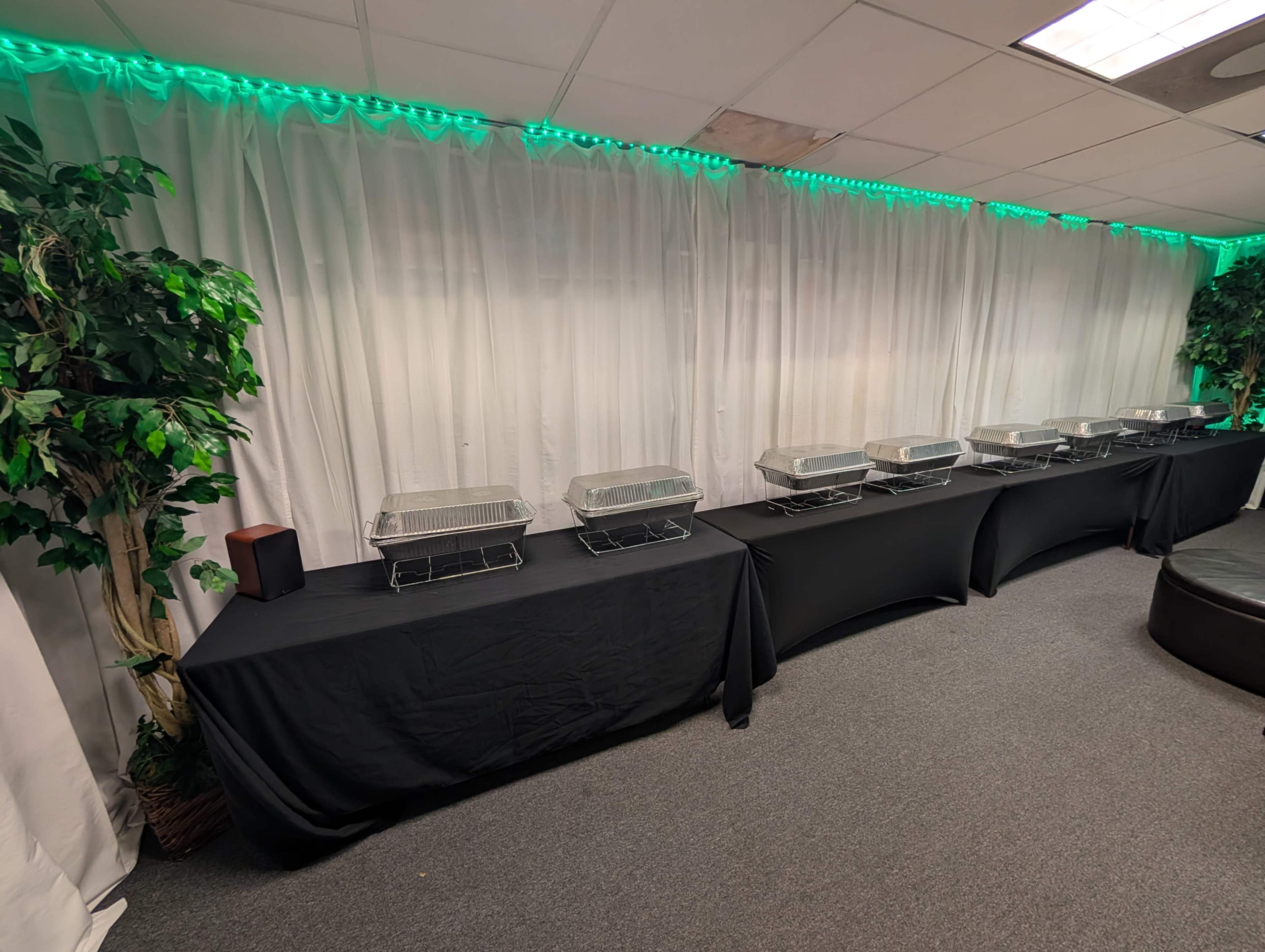 The image shows a banquet setup with several chafing dishes arranged on a long table covered by a black cloth, against a backdrop of white drapery and green lighting.