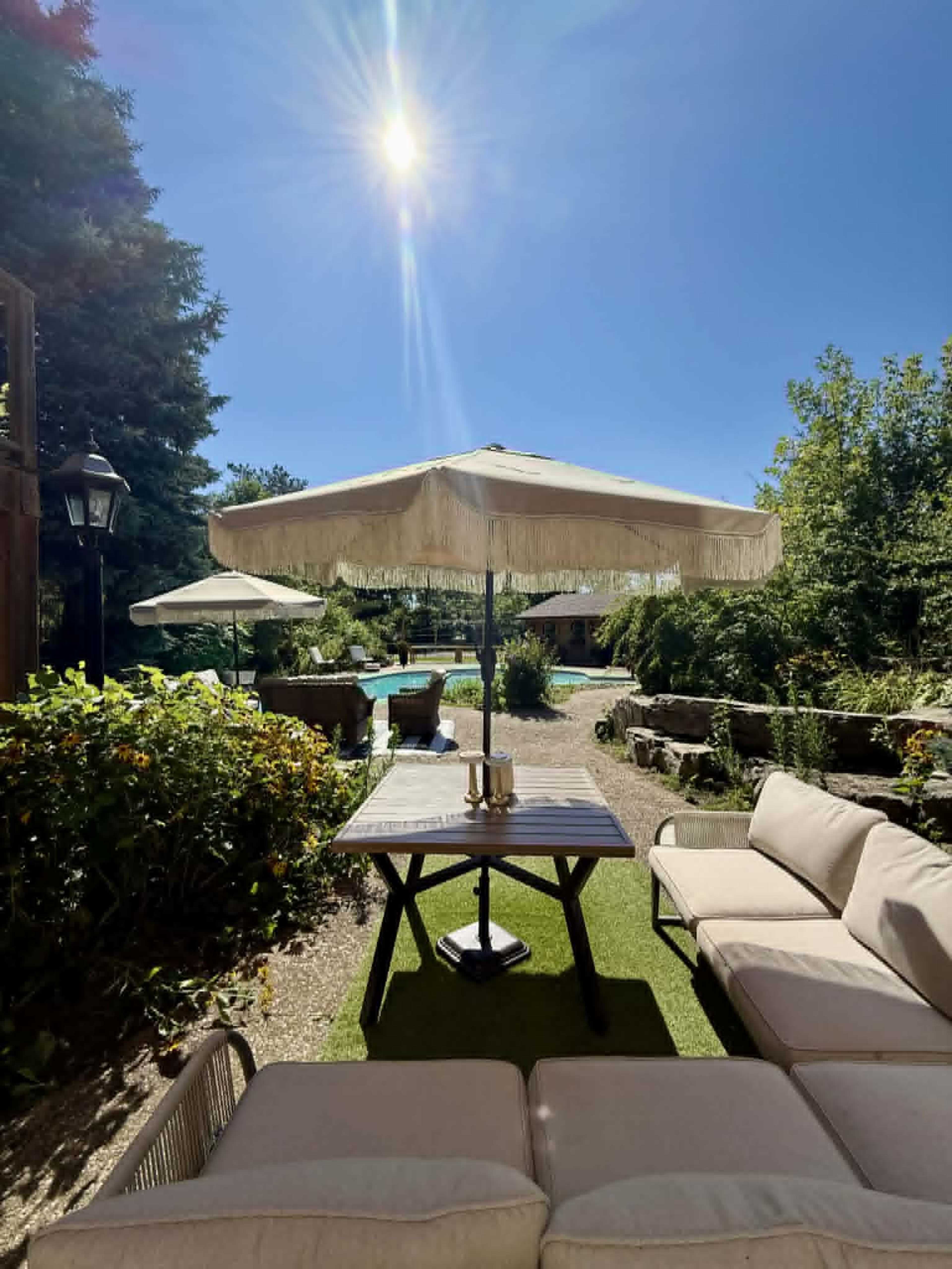 A cozy outdoor seating area with a table, shaded by a large umbrella, overlooks a pool and landscaped garden under a clear blue sky.