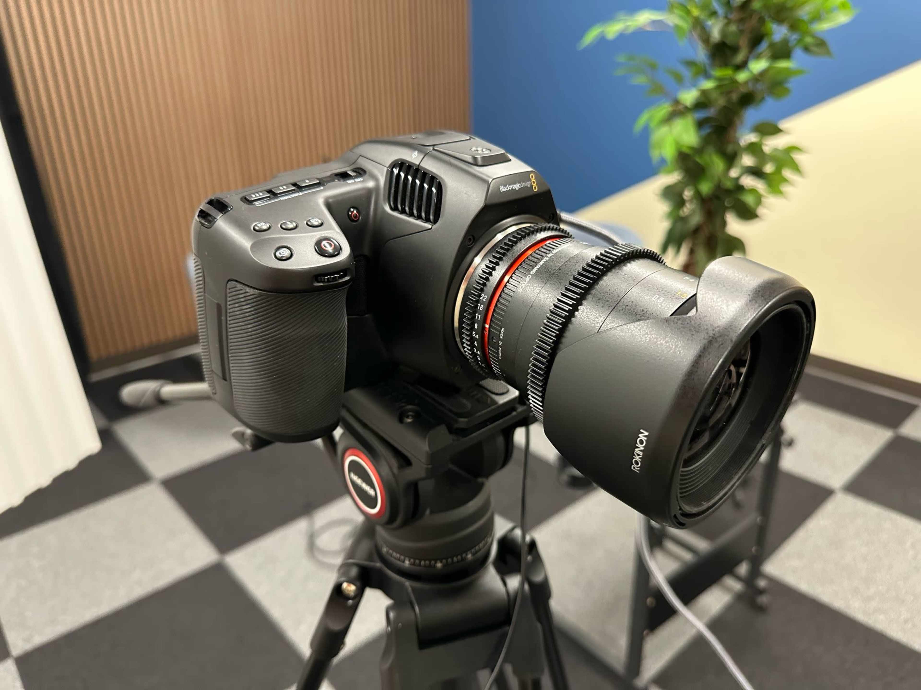 A digital camera is mounted on a tripod in a studio setting with patterned flooring and a potted plant in the background.