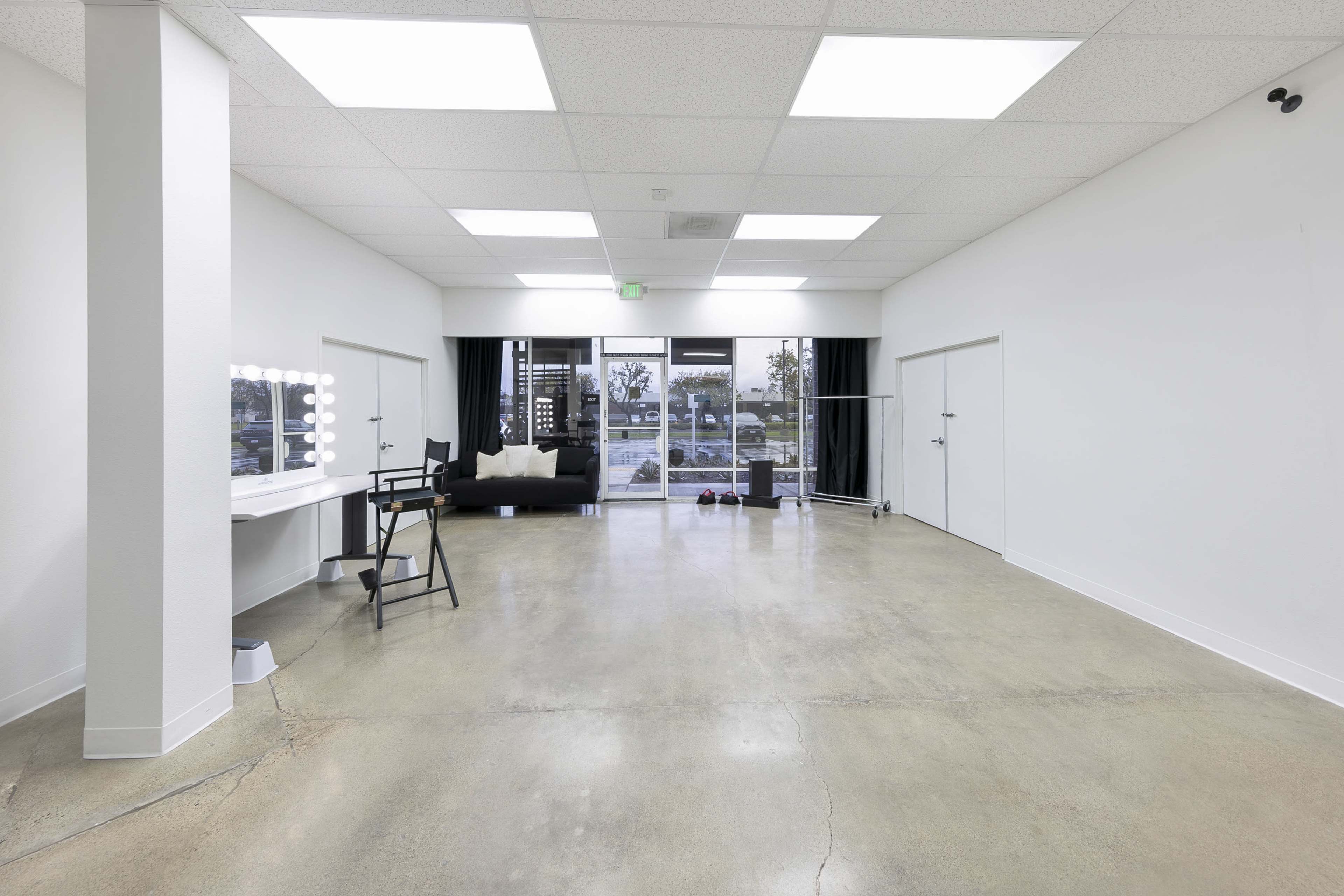 The image shows a spacious, well-lit studio with a polished concrete floor, a makeup vanity with lights, a black chair, and large glass doors leading to an outdoor area.