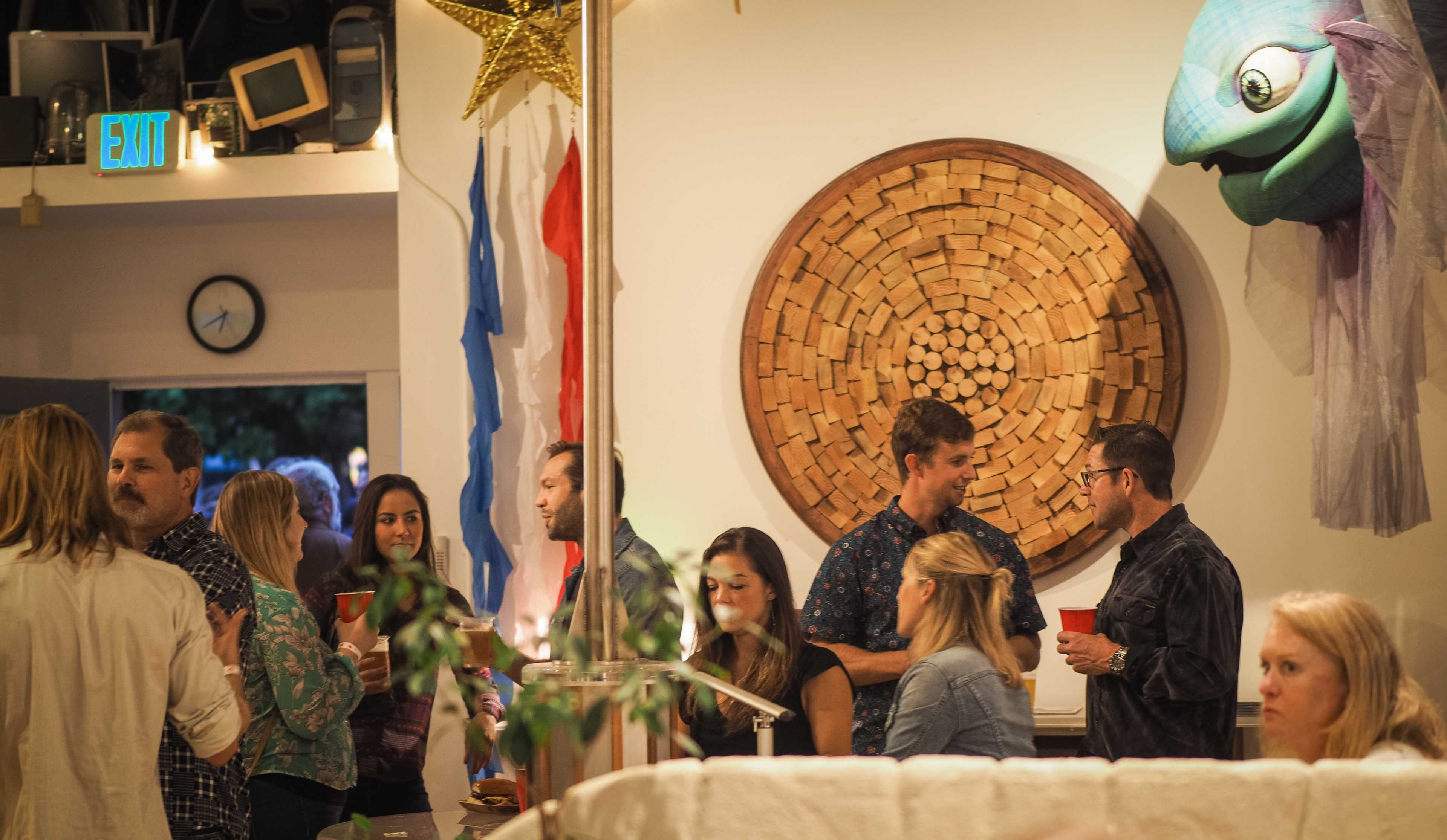 A group of people socialize at a gathering in a room decorated with a large circular wooden wall art and a colorful fish hanging from the ceiling.