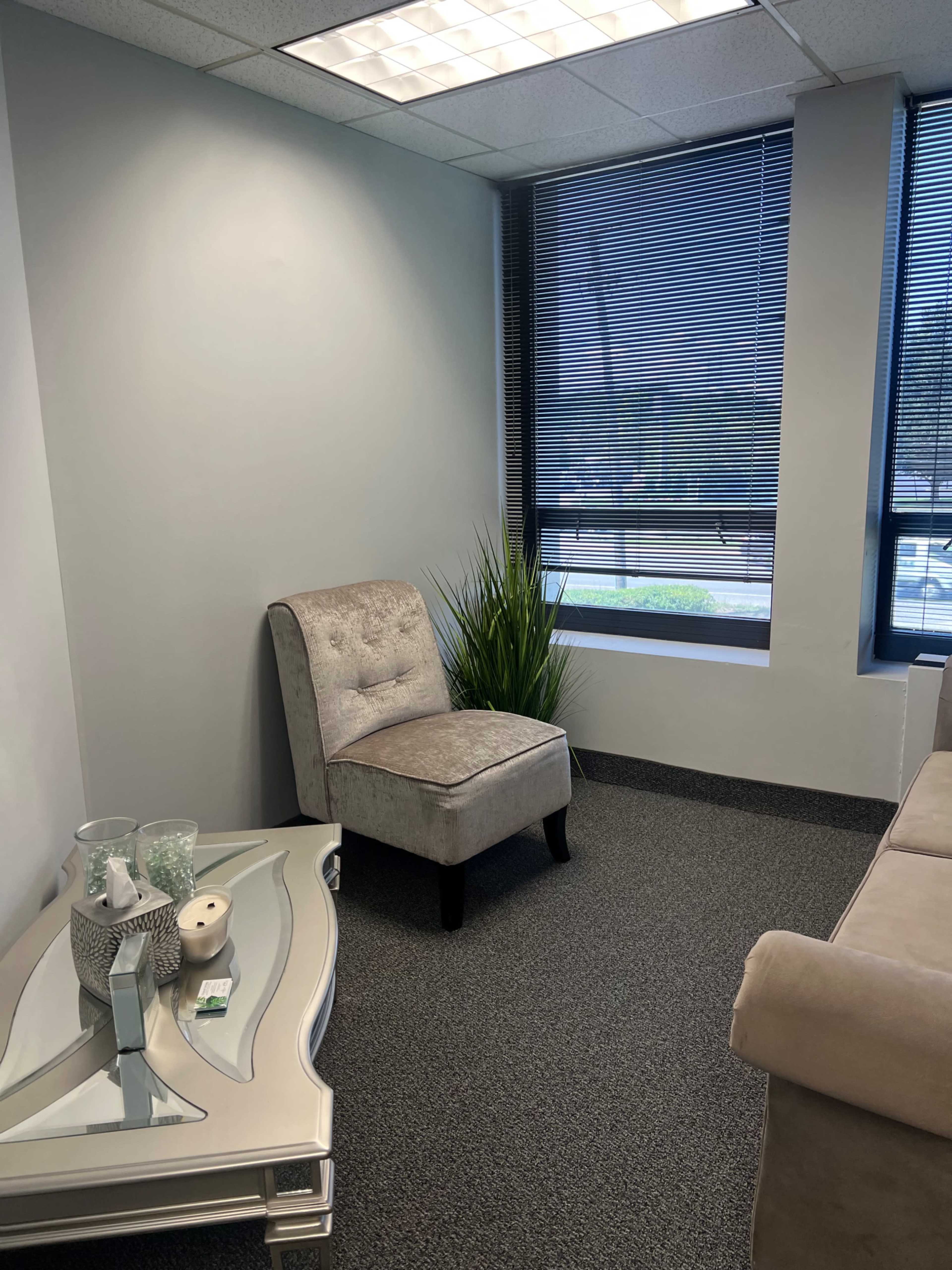 The image shows a small waiting area with a gray upholstered chair, a decorative plant, and a light-colored table next to a sofa, all set against a backdrop of large windows.