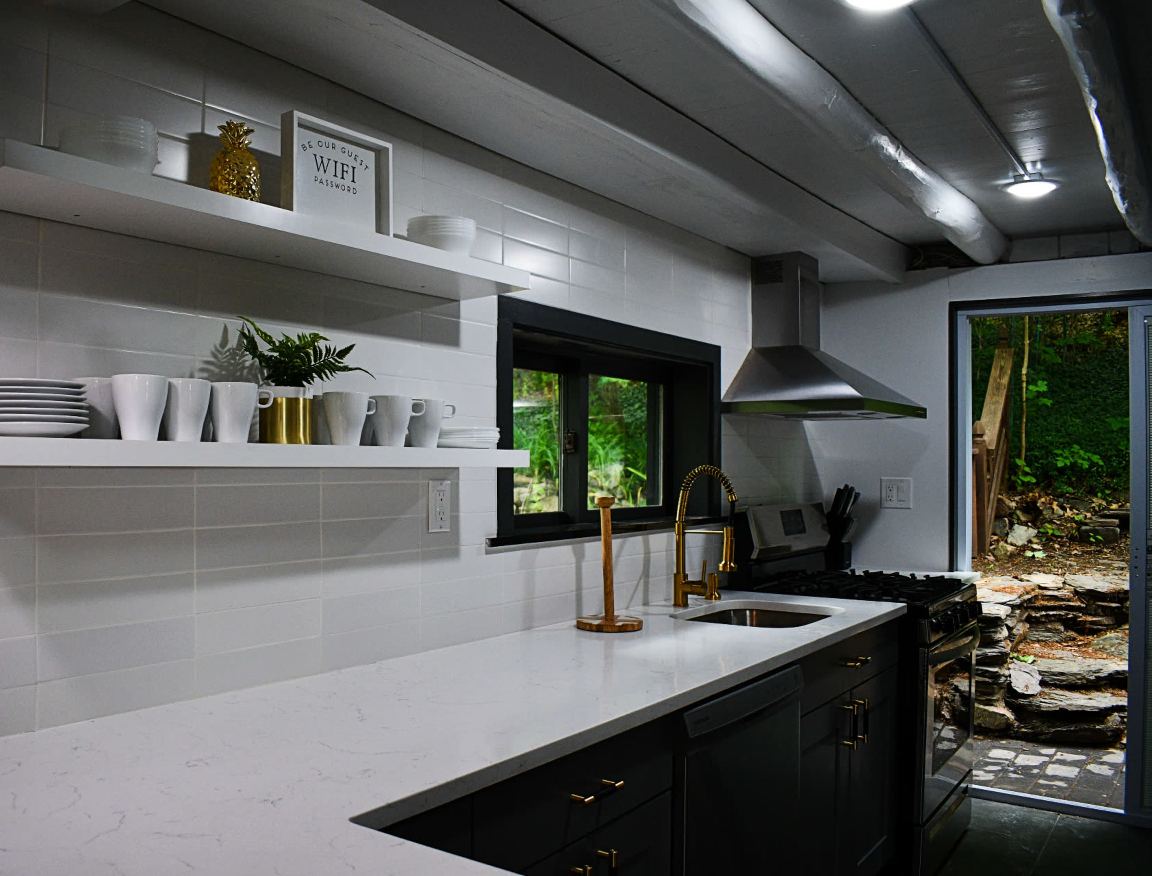 A modern kitchen with white tile walls, a large countertop, open shelves displaying dishware, and a view of an outdoor stone area through a glass door.
