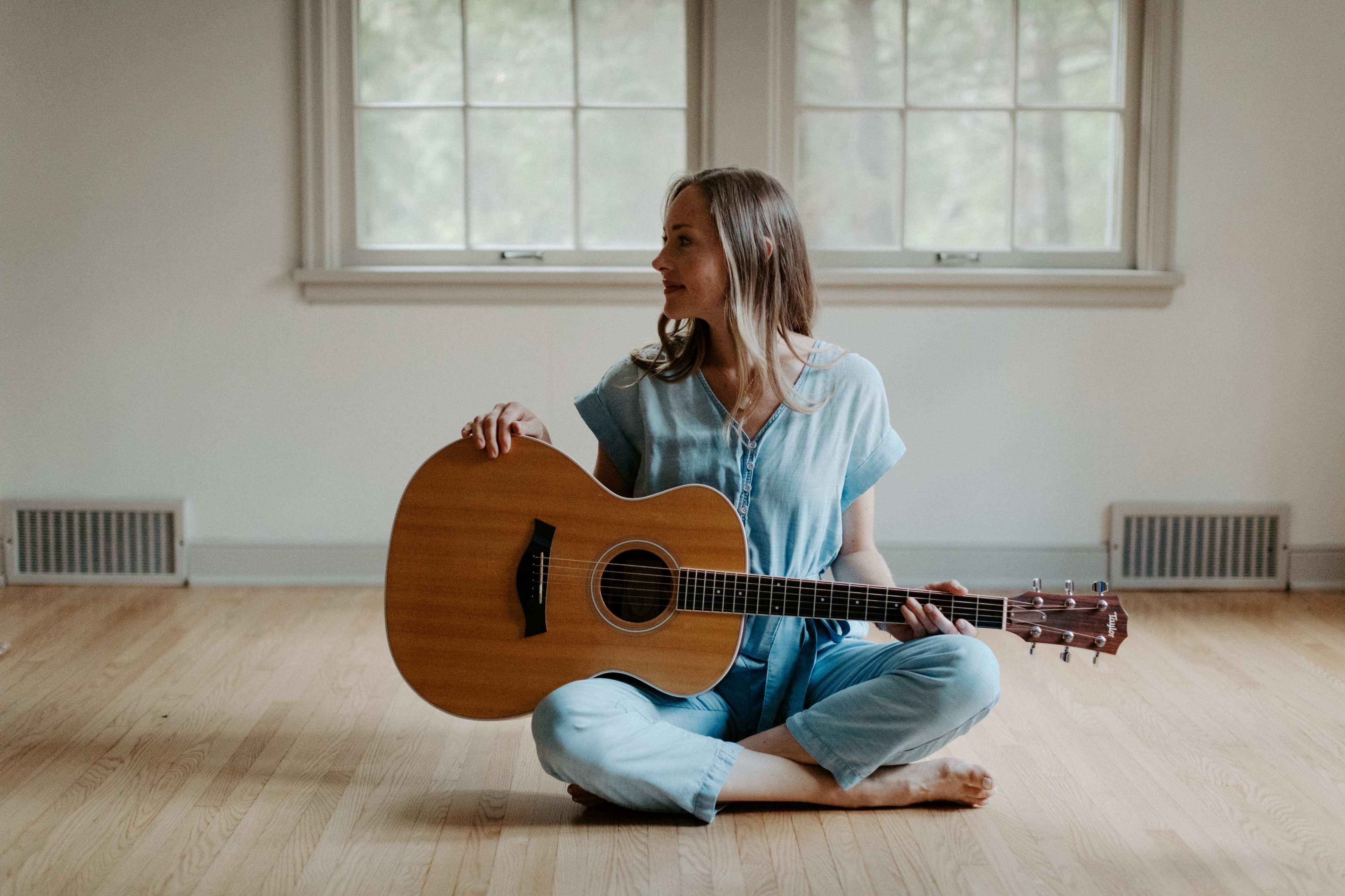 A woman sits cross-legged on a wooden floor, holding an acoustic guitar, with two windows in the background.