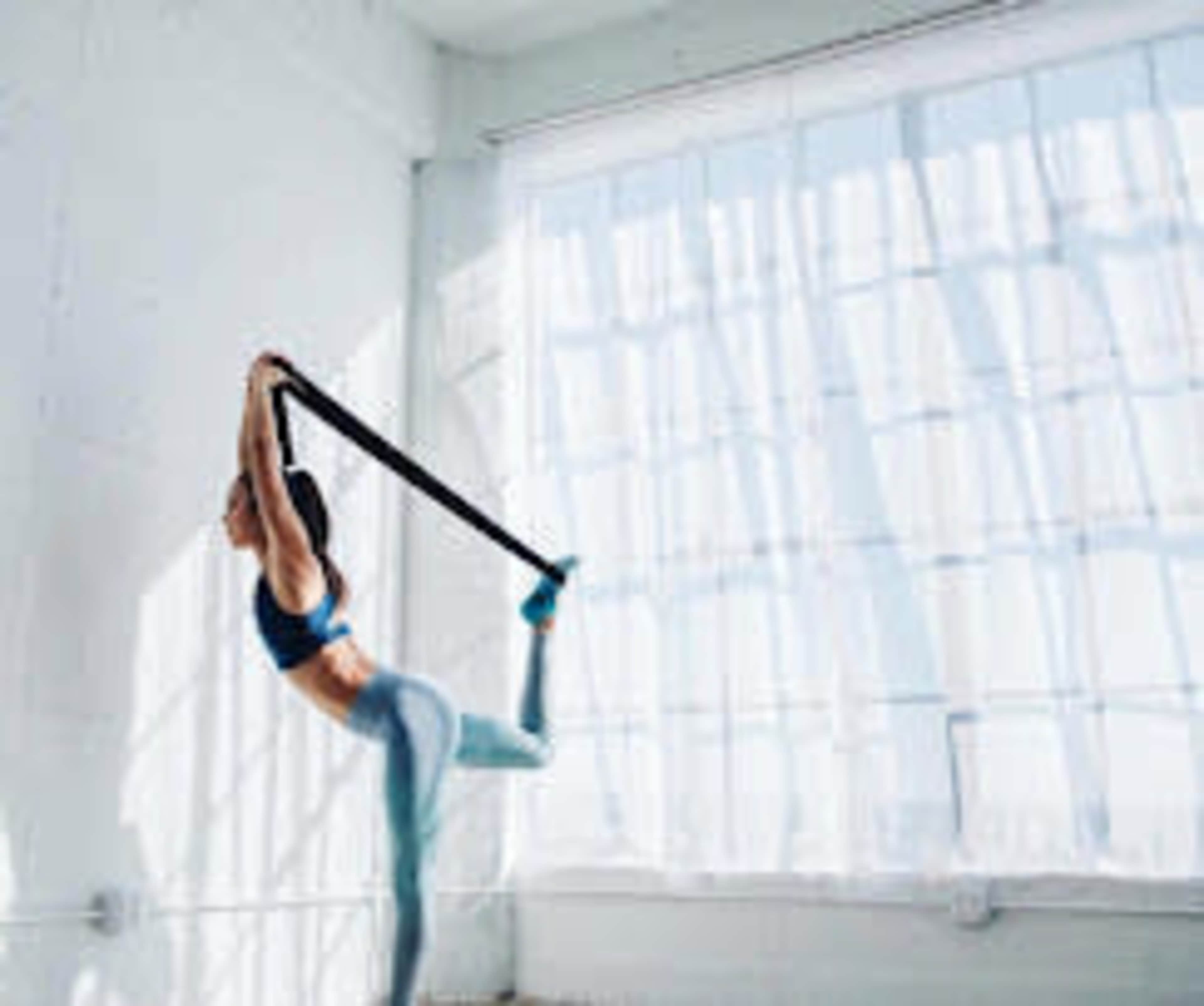A woman performs a stretching exercise using a resistance band in a brightly lit room with large windows.