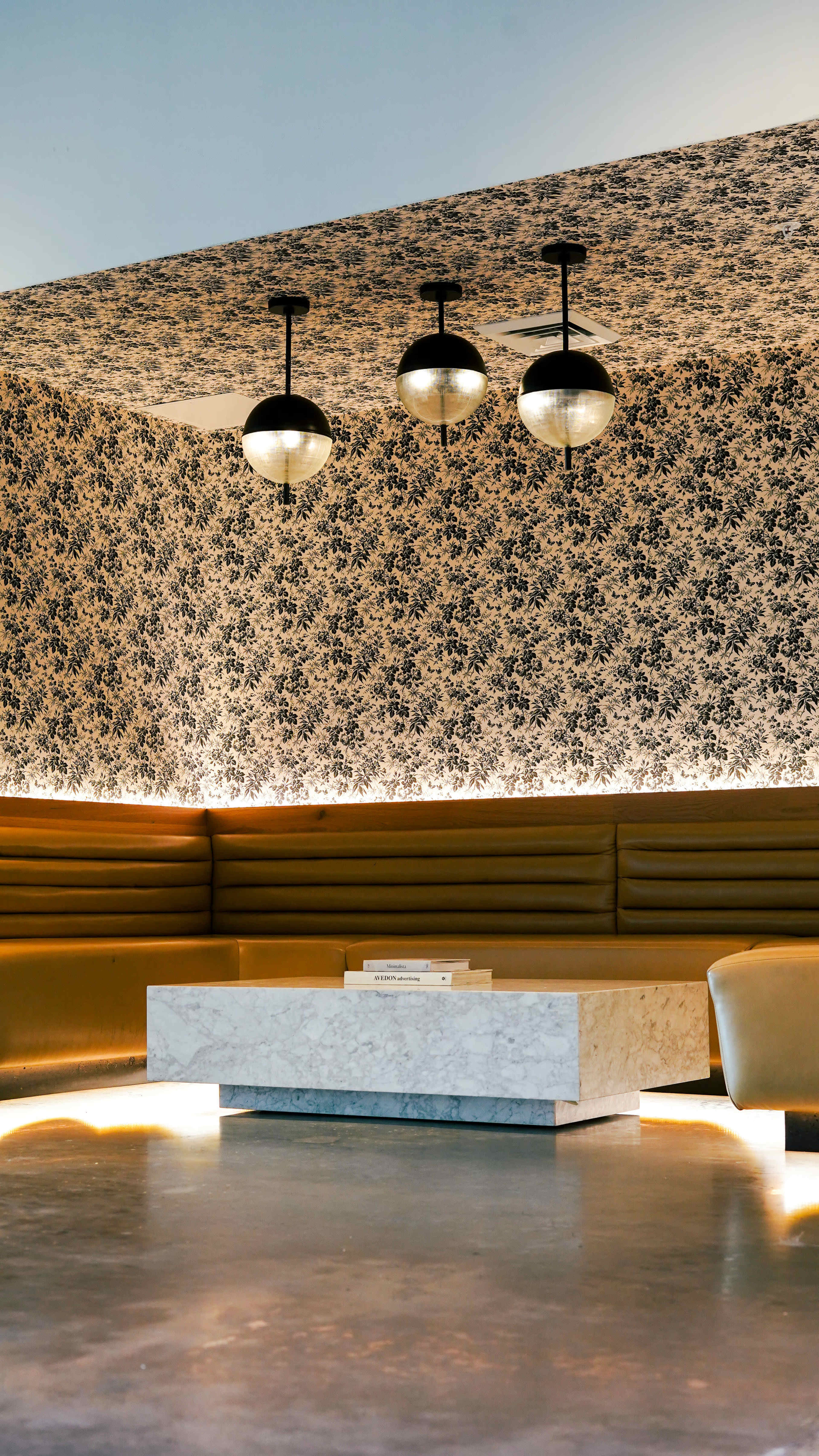 The image shows a modern interior space with a patterned wallpaper, seating along the wall, and three pendant lights hanging above a marble coffee table.