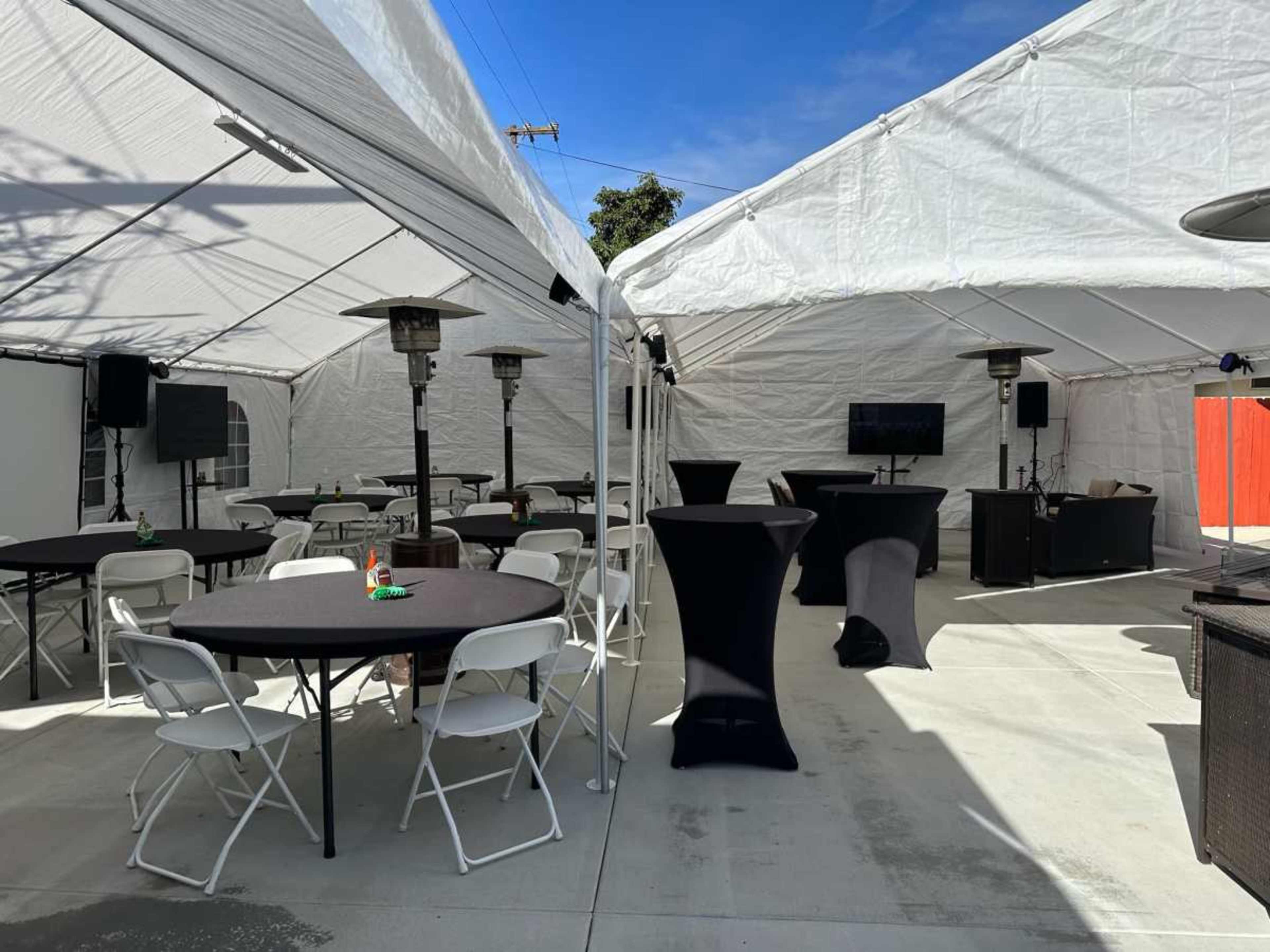 A large outdoor tent is set up with several high tables, chairs, and heating lamps, providing a space for gatherings.