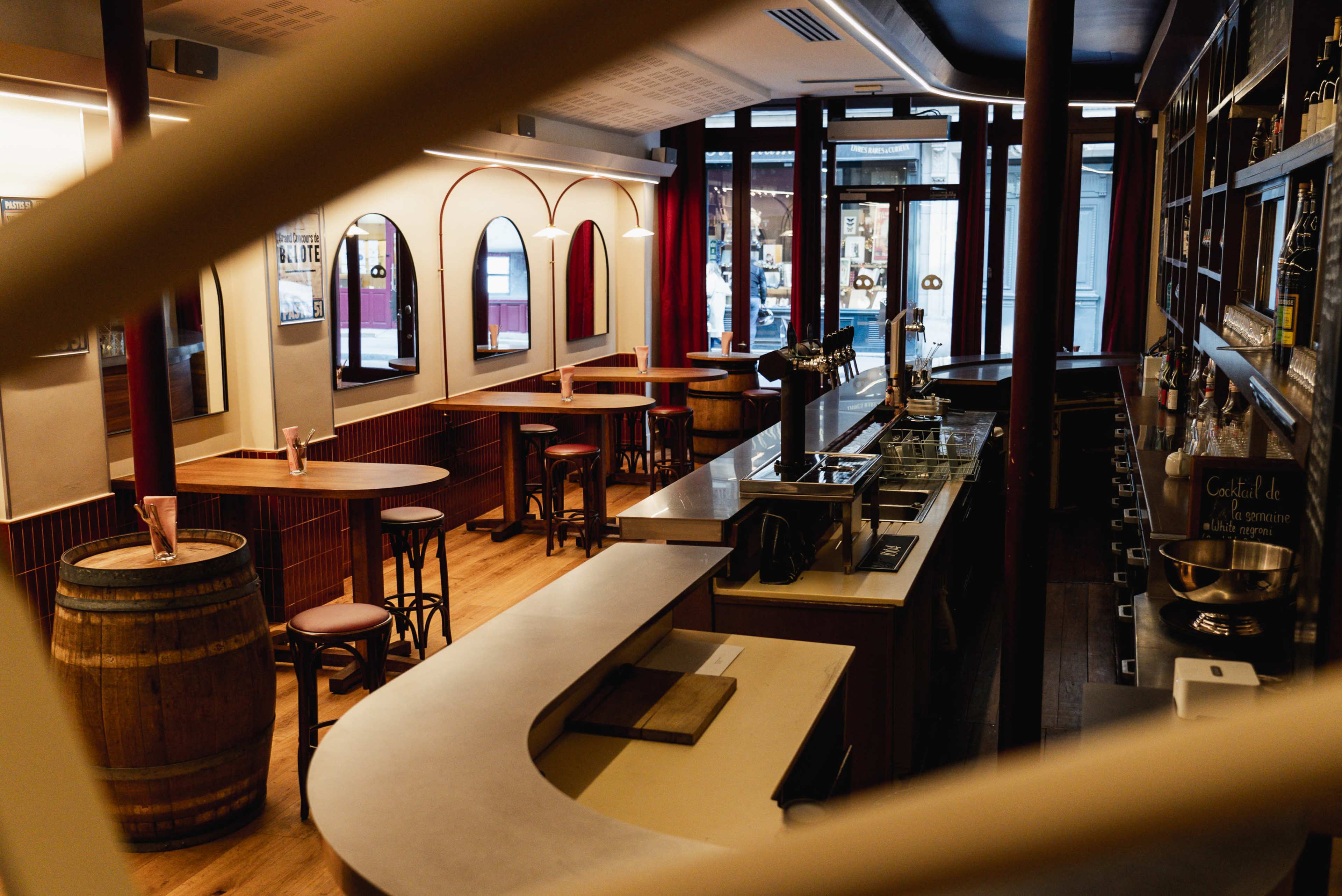 The image shows an empty bar interior with wooden tables, high stools, and a counter, framed by large windows and decorative mirrors.