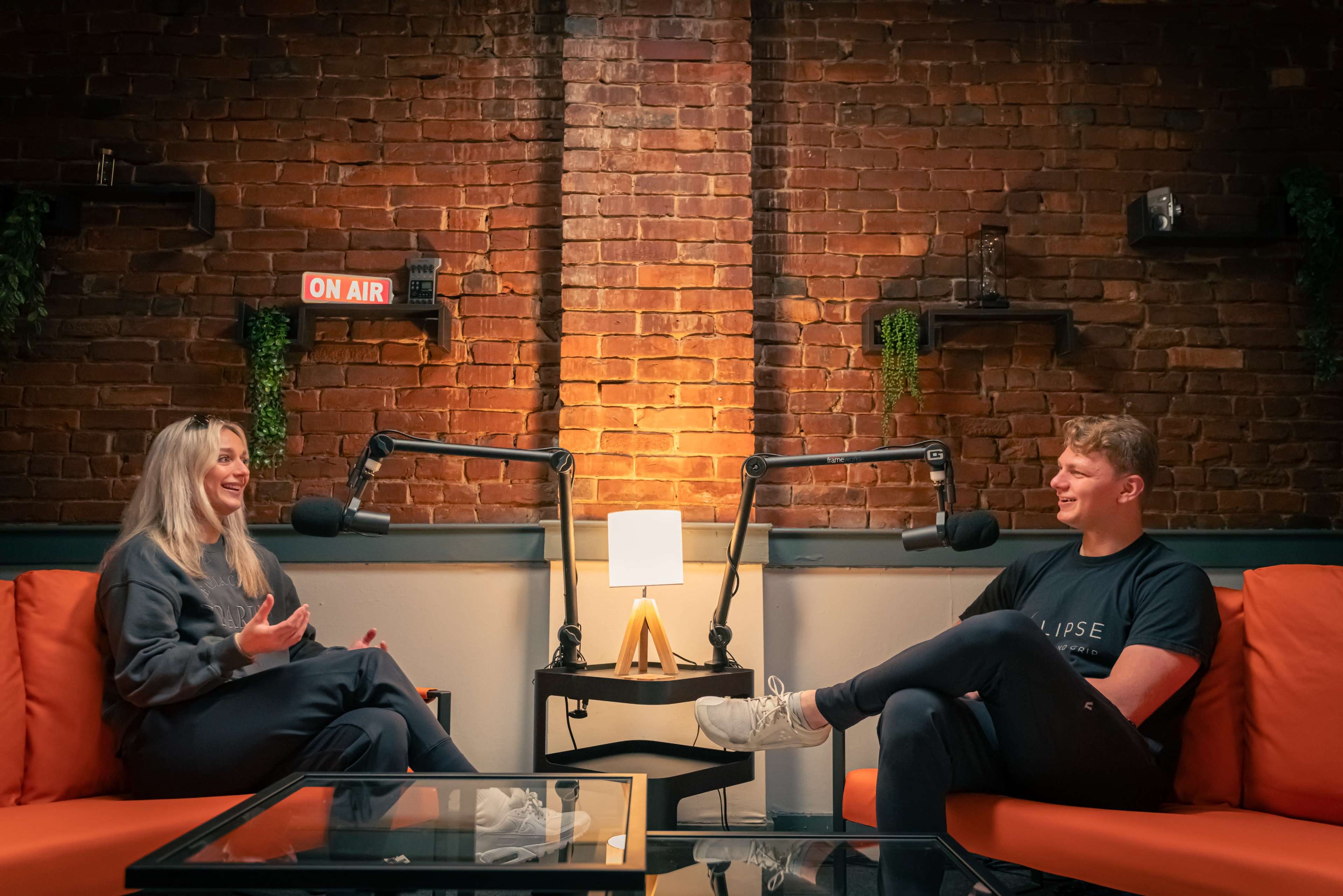 Two individuals are seated in a cozy studio setting with orange couches, engaged in conversation while microphones are positioned in front of them, and a lamp illuminates the space.