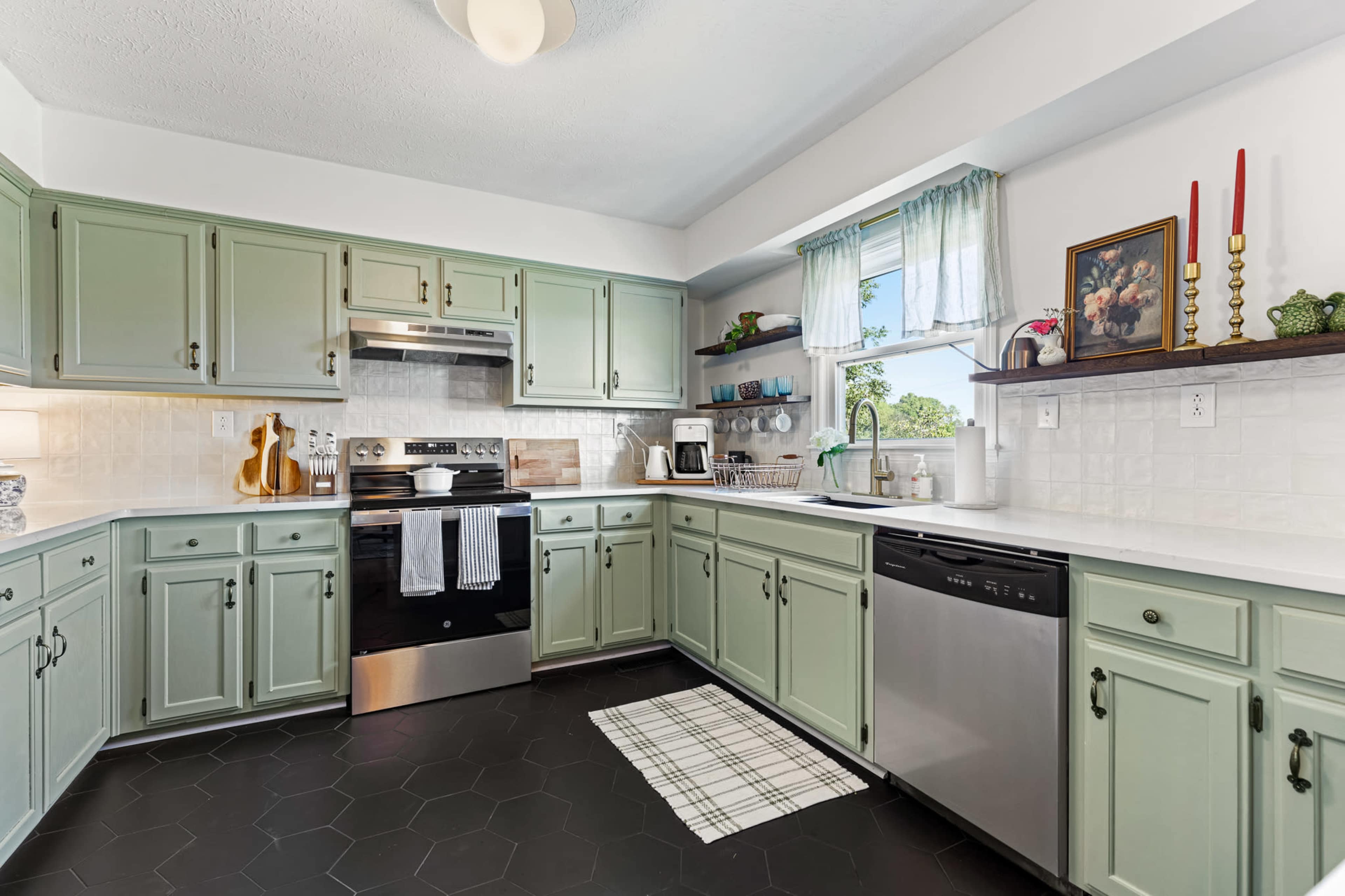 The image shows a modern kitchen with green cabinets, black appliances, and a window above the sink.
