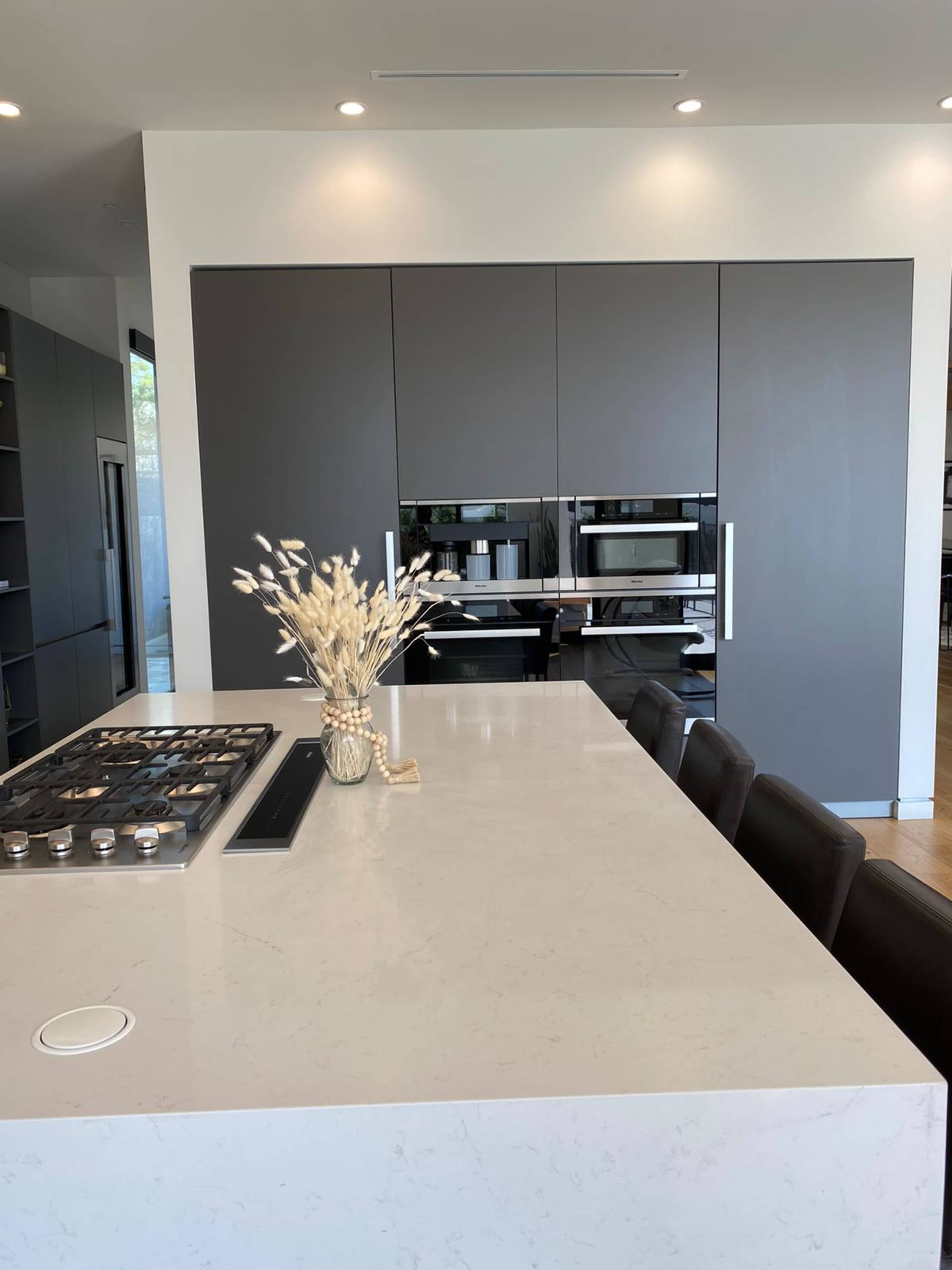 A modern kitchen features a large white countertop with a stovetop and a decorative vase of dried flowers, set against dark cabinetry and dual wall ovens.